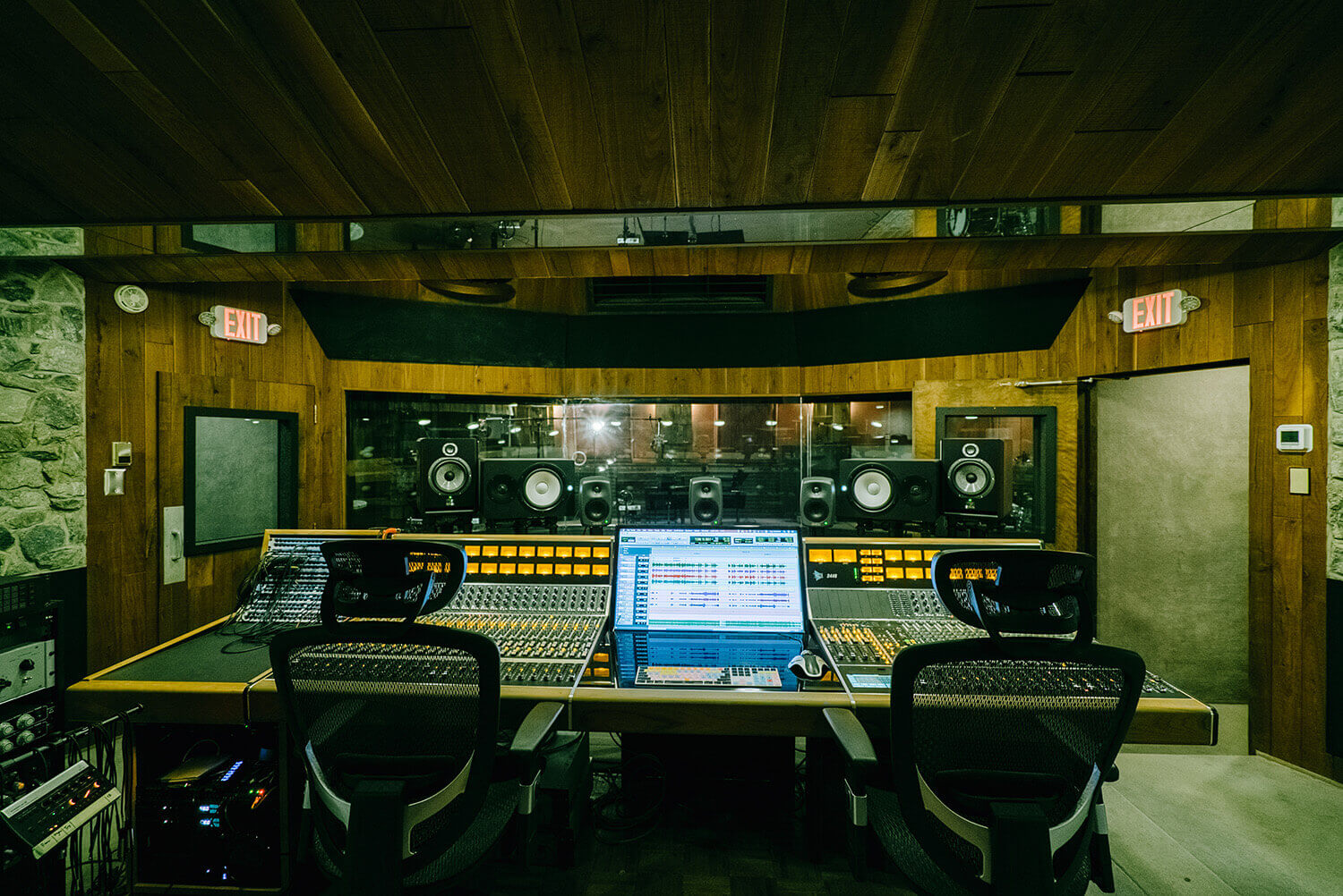 Audio mixing studio with two chairs at a large console, computer screen showing audio tracks, speakers on shelves, wood walls and ceiling, and a glass window between rooms.