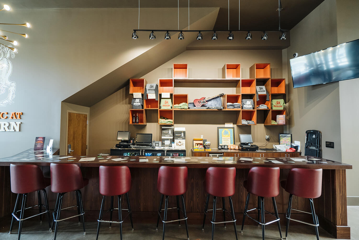 Modern bar with a long wooden counter, six red cushioned stools, shelves with merchandise, books, decor, and a TV on the right wall.