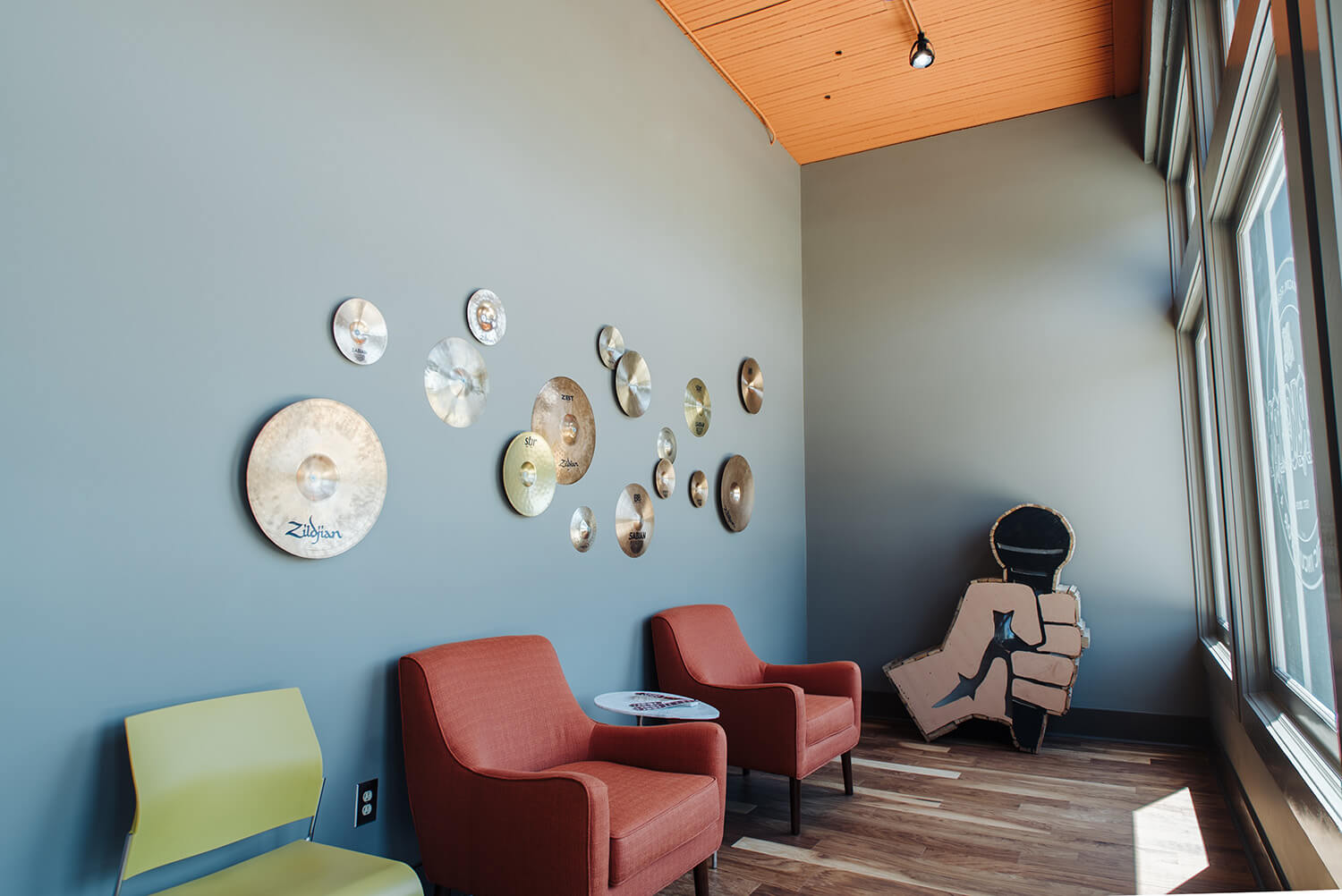 Cozy lounge with two orange armchairs, a green chair, cymbals on a blue-gray wall, large window, and fist art on wood floor.