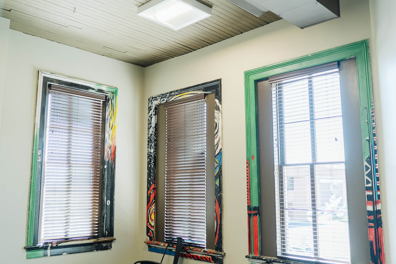 Three windows with wooden blinds in a corner, colorful abstract frames, sunlight streaming through, part of a chair below.