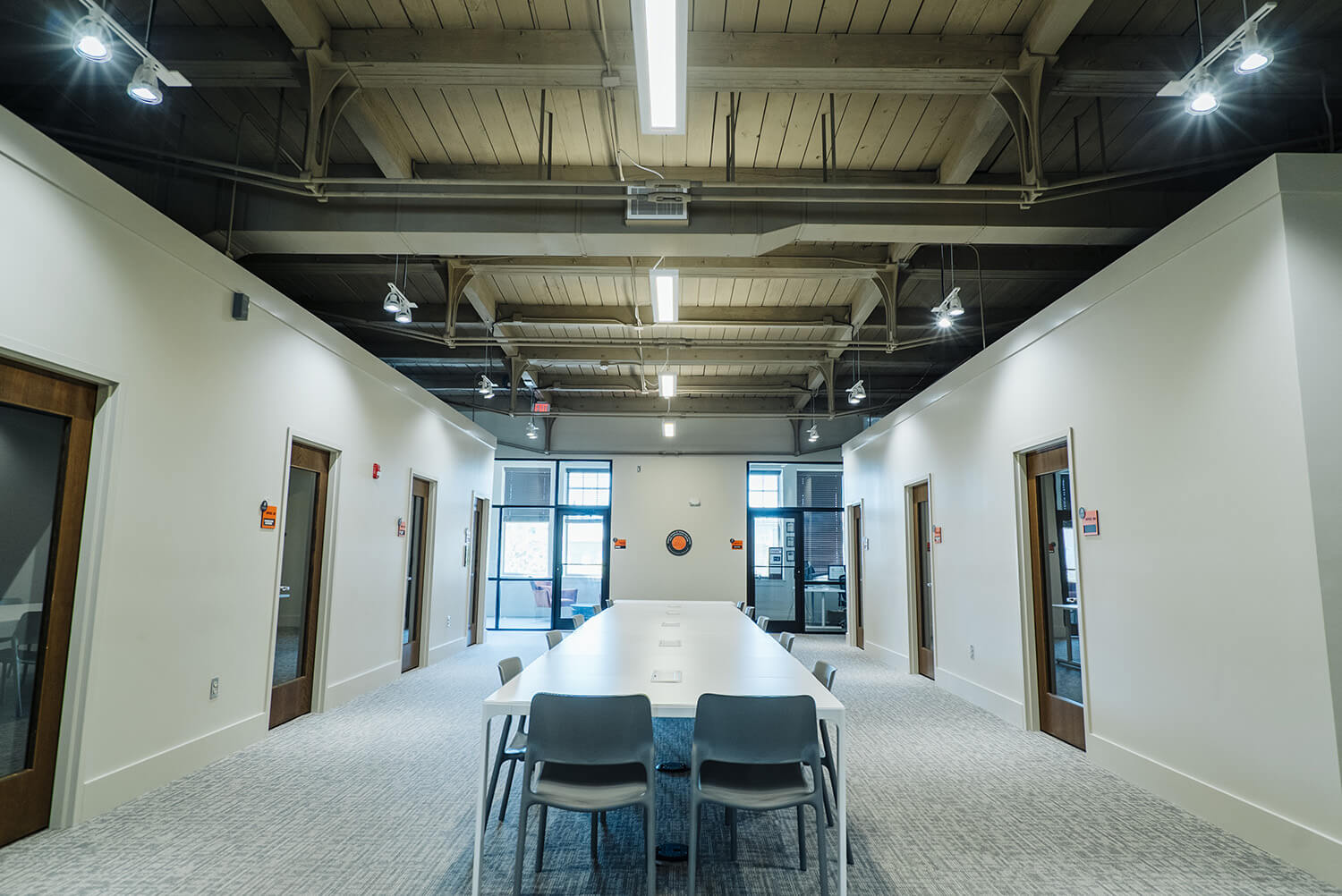 Modern conference room with a long white table, several chairs, exposed beam ceiling, bright lighting, and large windows at the end.