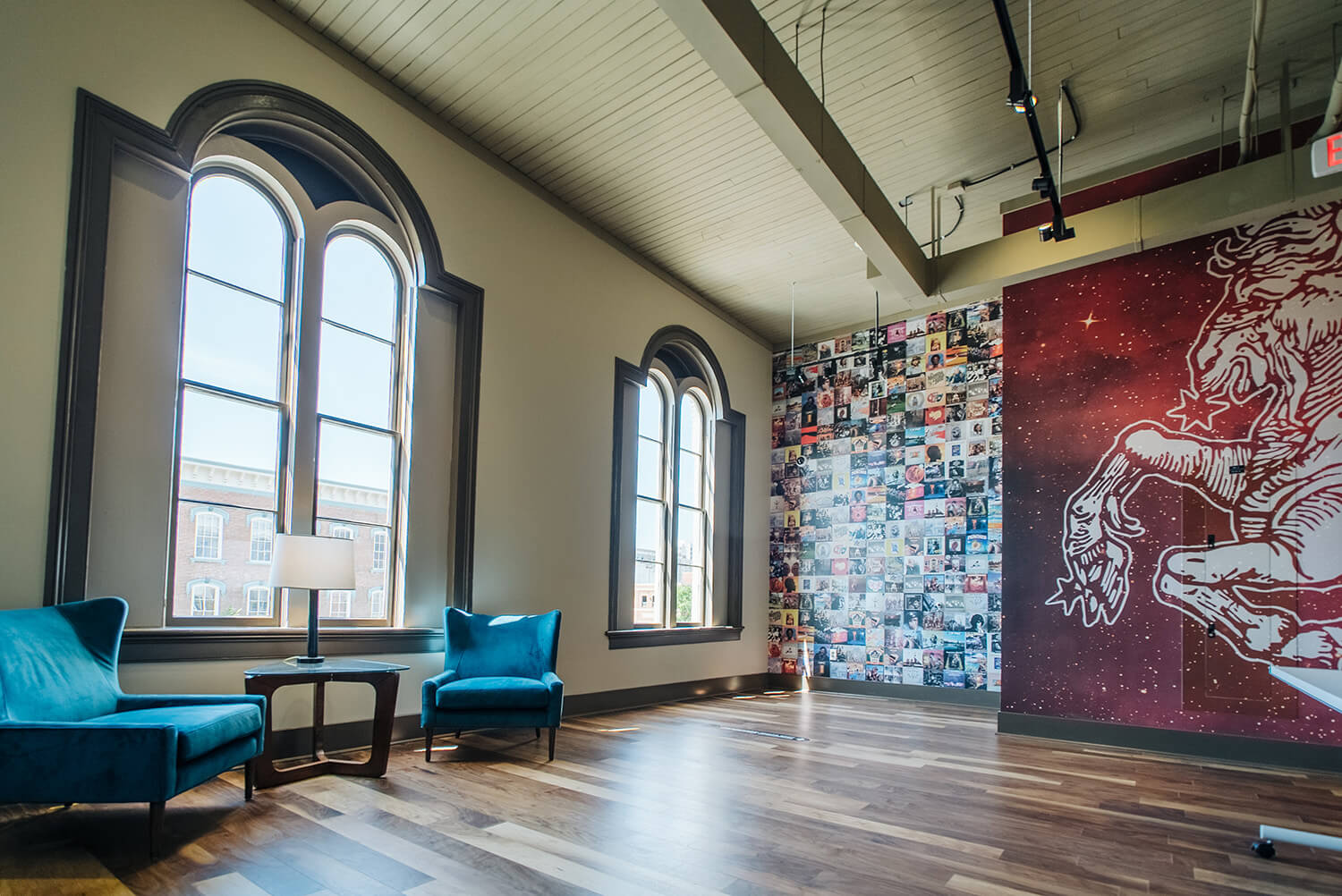 Modern lounge with arched windows, two blue chairs, feature wall of album covers and unicorn mural, hardwood floors, high ceilings.