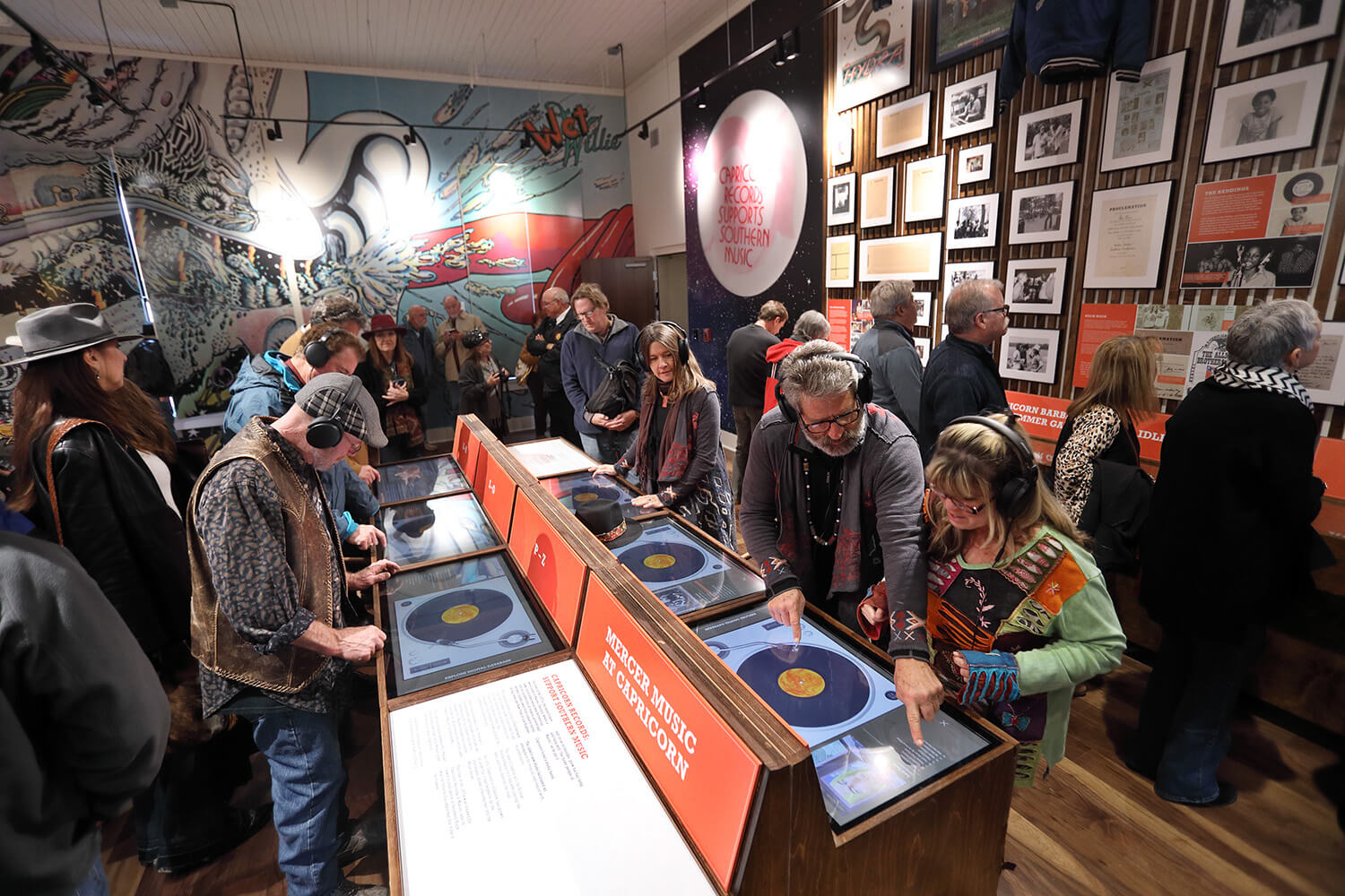 People explore interactive music exhibits in a museum, using headphones and reading displays. Colorful murals fill the background.
