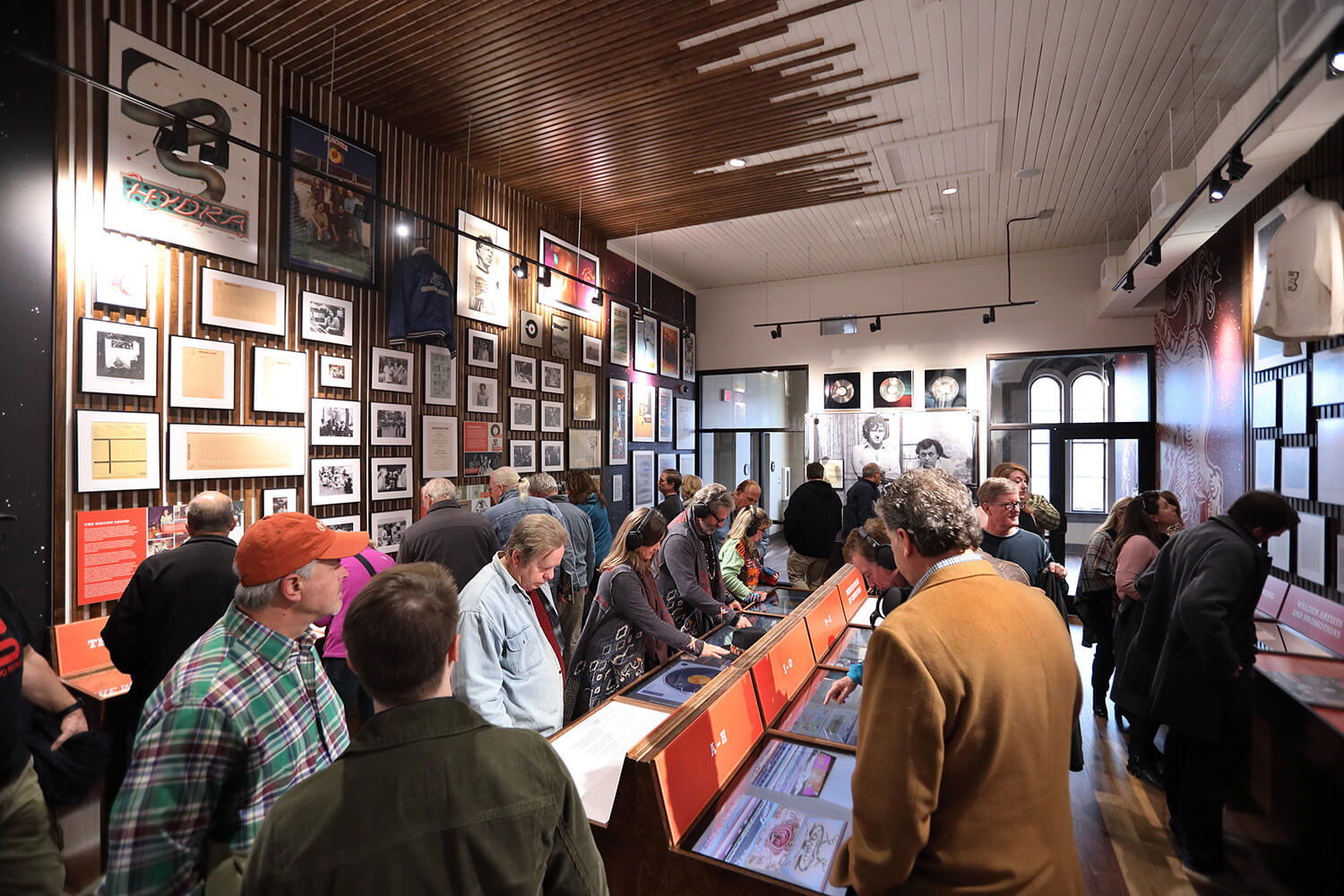 A crowd explores interactive exhibits and framed art in a museum gallery with wood ceilings, photo-filled walls, and memorabilia displays.