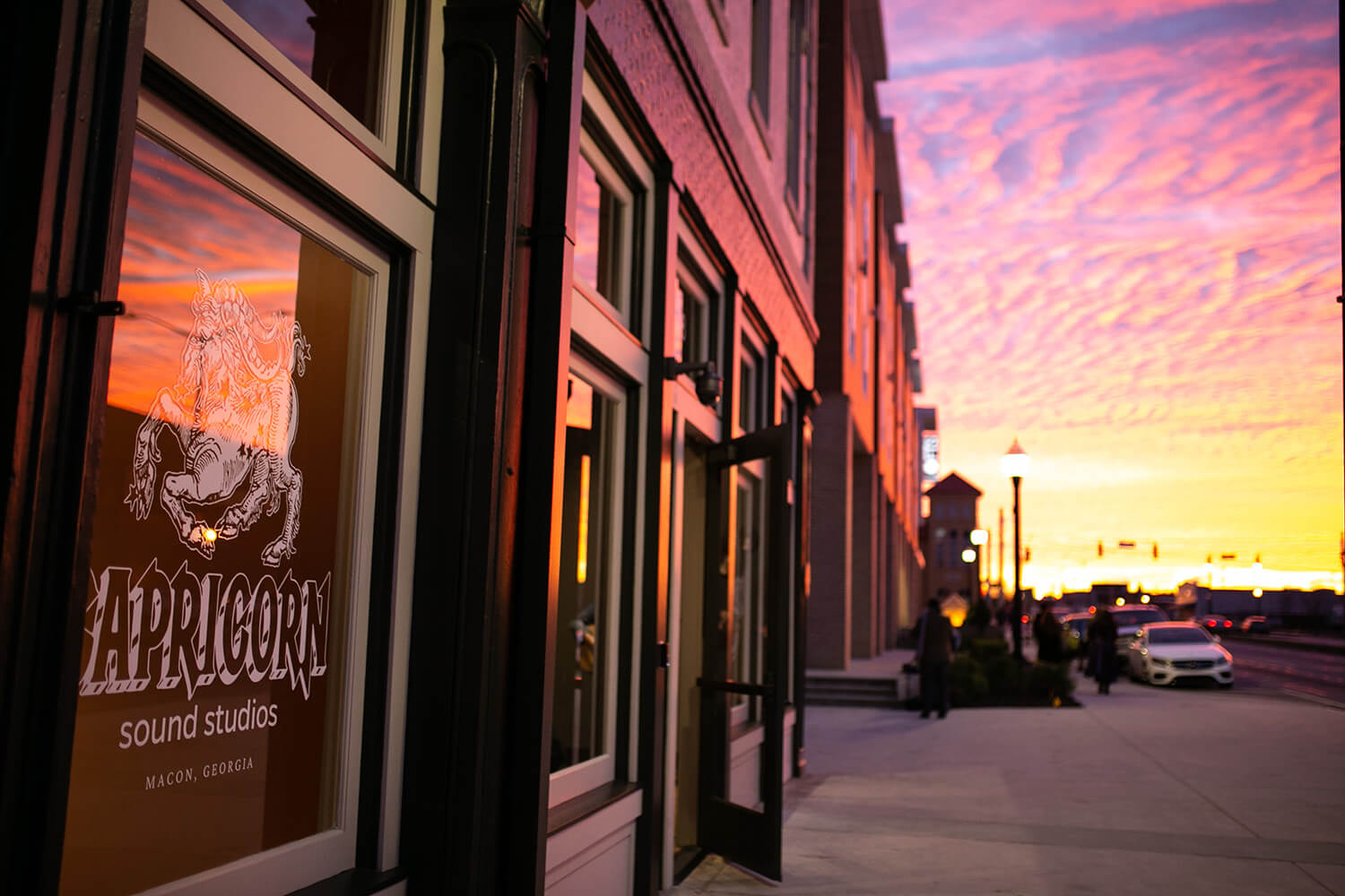 A vivid sunset reflects on the windows of Capricorn Sound Studios in Macon, Georgia, with people and cars along the lit sidewalk.