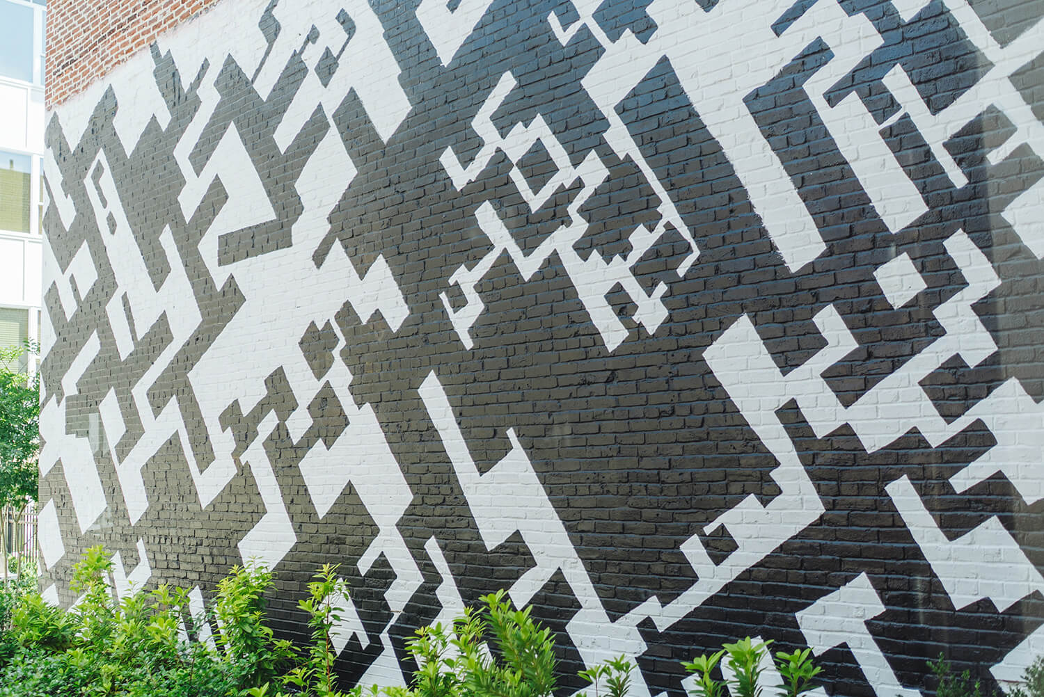 A brick wall with an abstract black and white geometric pattern of sharp shapes, and green plants growing at the base of the wall.