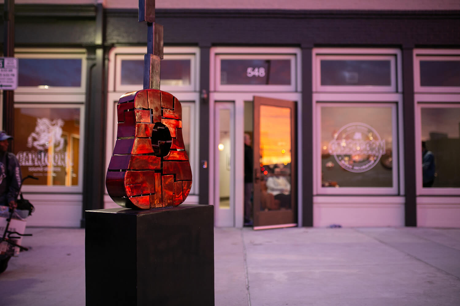 A fragmented red guitar sculpture sits on a black pedestal outside a building with large windows; the address 548 is above the door.