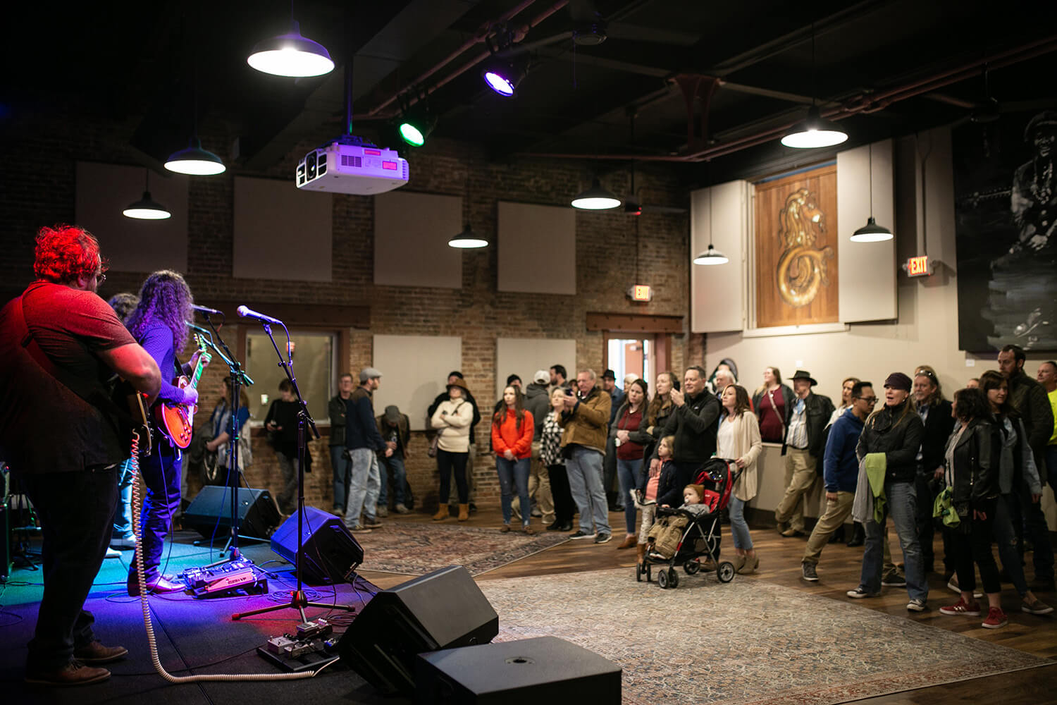 Musicians play guitars onstage in an indoor venue. The audience stands, some in winter clothes. Art decorates brick walls. Stage lights shine.