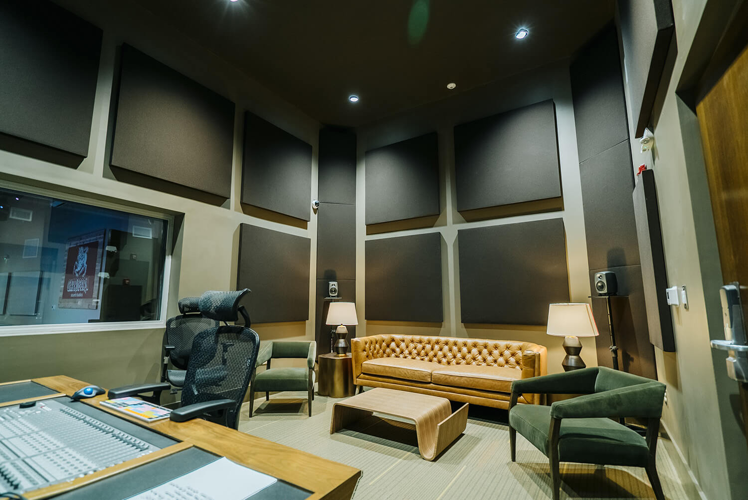 Modern recording studio with soundproof panels, tan leather sofa, green chairs, mixing console, and window; warm table lamp lighting.
