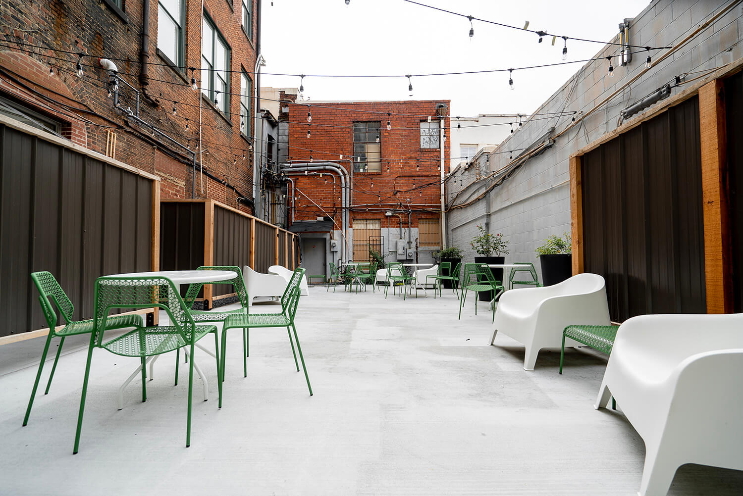 An outdoor patio features green metal tables, chairs, and white plastic loungers. String lights hang overhead. The patio is empty.