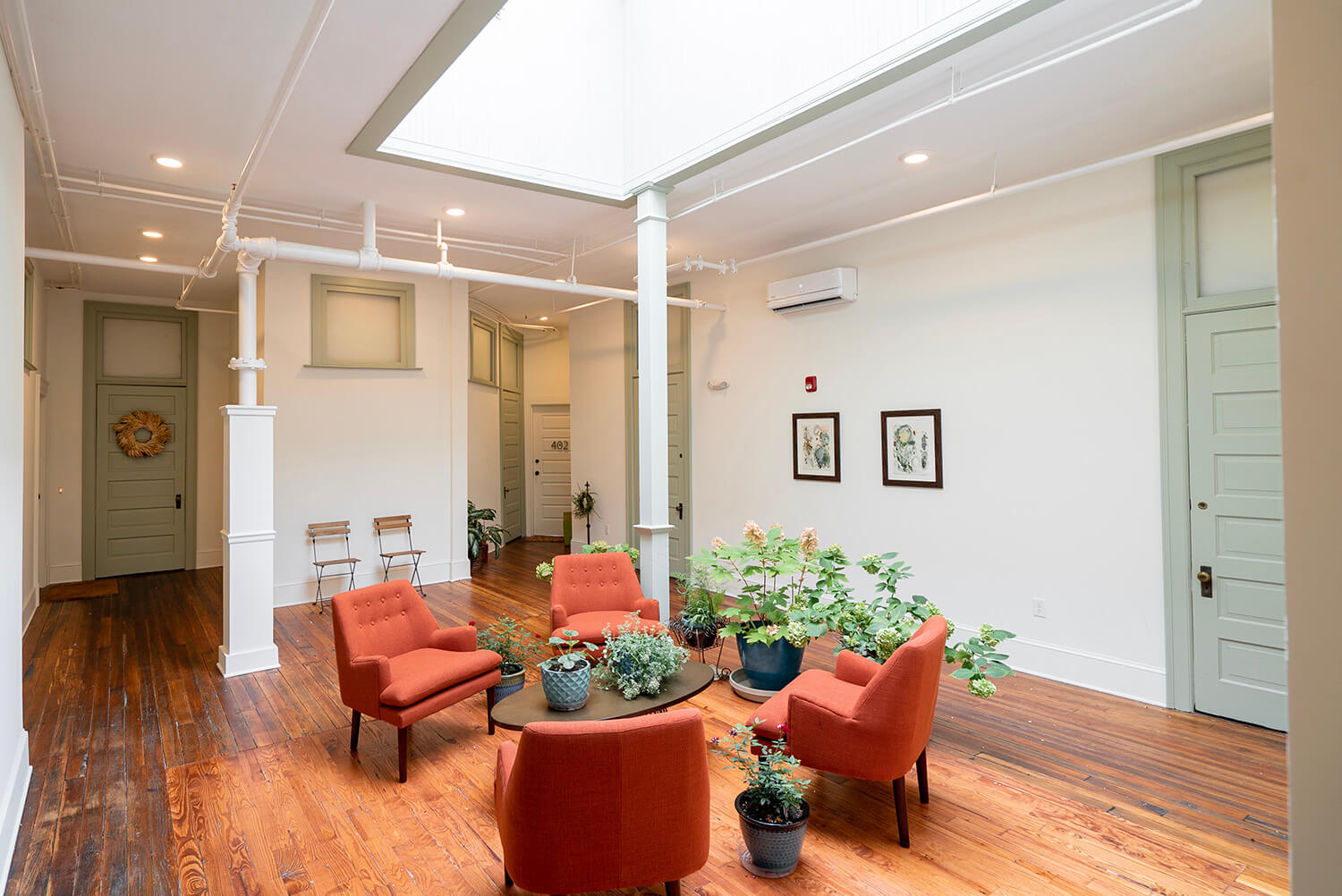 Bright, airy living room with hardwood floors, four orange armchairs around a table with plants, skylight, pale green doors, framed art.