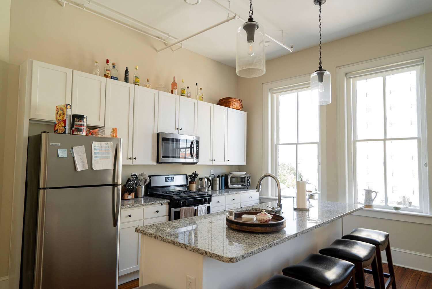 Modern kitchen with white cabinets, stainless appliances, granite counters, four black island stools, and sunlight from two windows.