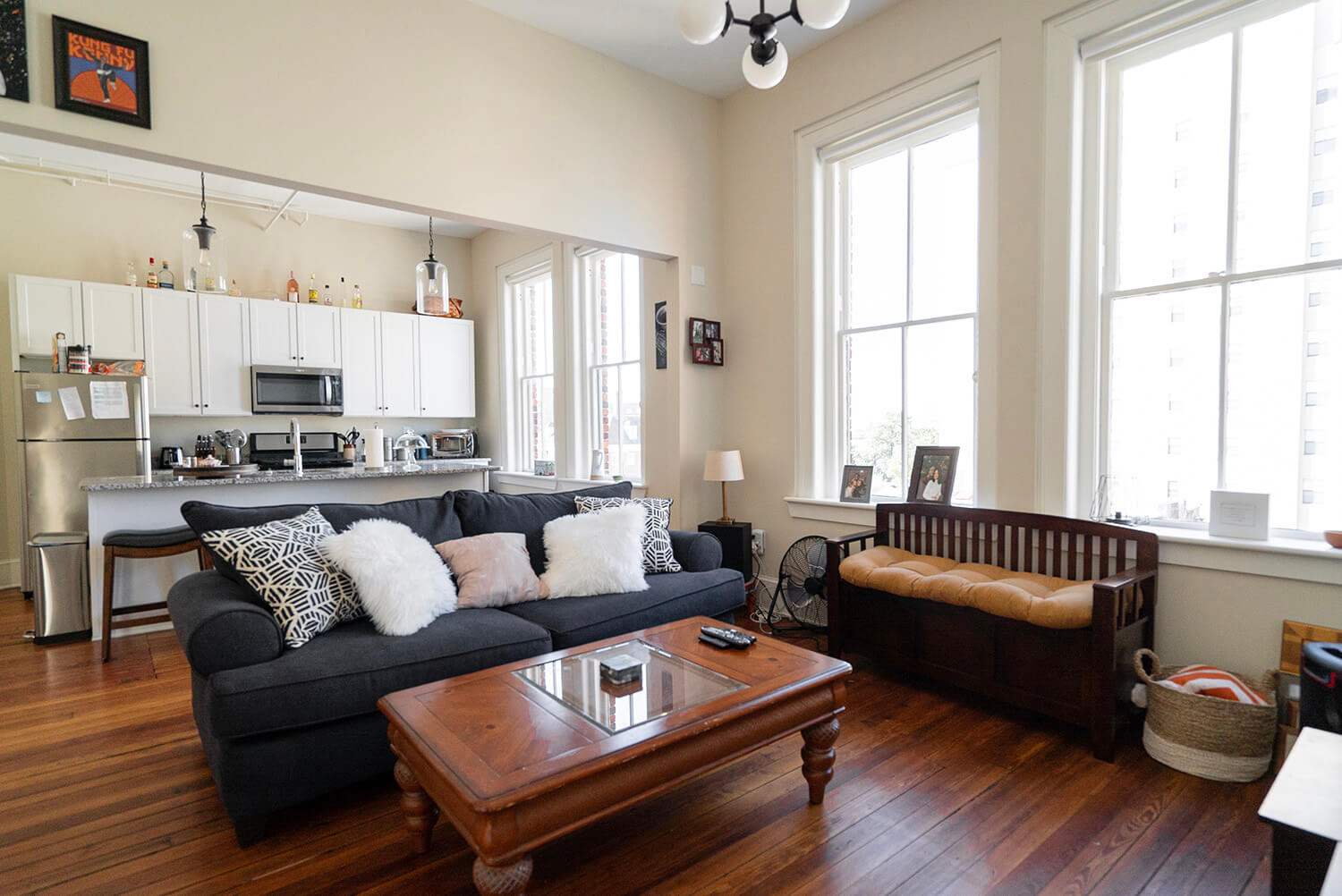 Cozy living room with dark sofa, fluffy pillows, wooden coffee table, big windows. Kitchen with white cabinets and hardwood floors.