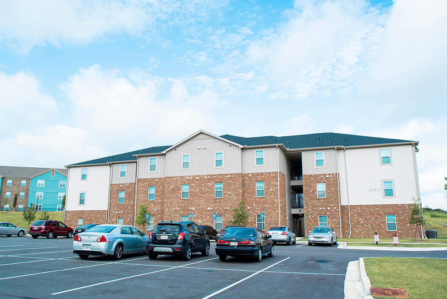Three-story apartment building with brick and light siding; several cars are parked in a mostly empty lot under partly cloudy skies.