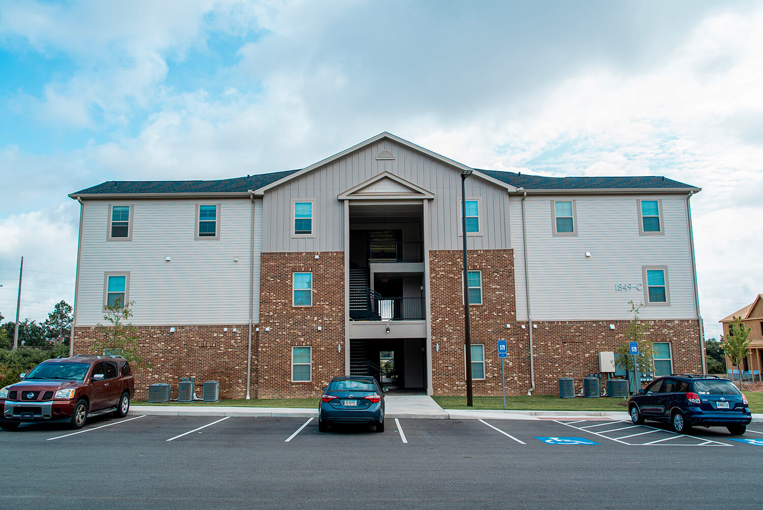 Three-story apartment building with brick and light siding, stairs at entrance, three cars in lot with accessible spaces on a cloudy day.