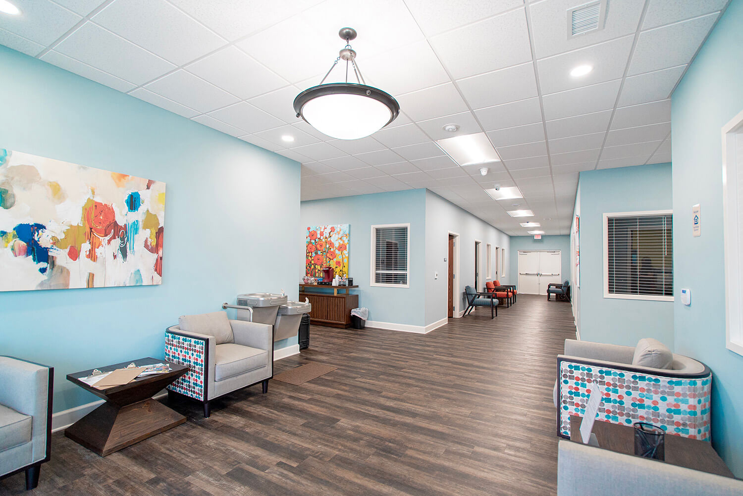 Bright modern waiting area with light blue walls, abstract art, patterned gray chairs, dark wood floors, and a well-lit hallway.