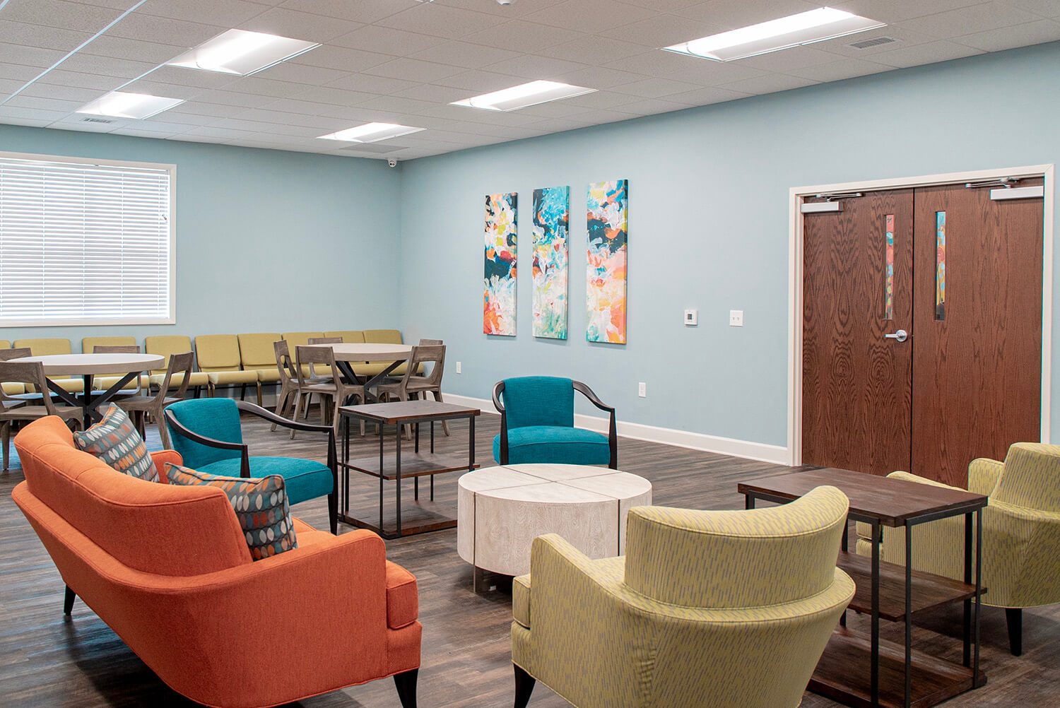 Modern waiting room with orange, blue, and yellow chairs, several tables, abstract art on pale blue walls, large window, and bright lighting.