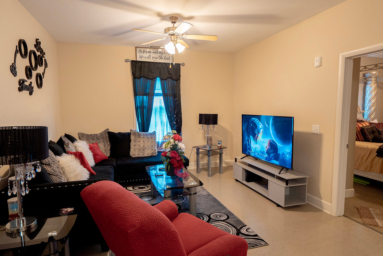 Cozy living room with red armchair, black sectional sofa, glass coffee table with flowers, TV showing sci-fi, bedroom visible.
