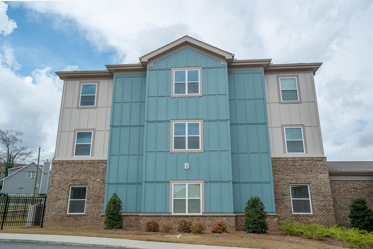 Three-story apartment building with beige siding, blue panels, brown brick base, six windows, a white B above entrance, and shrubs in front.