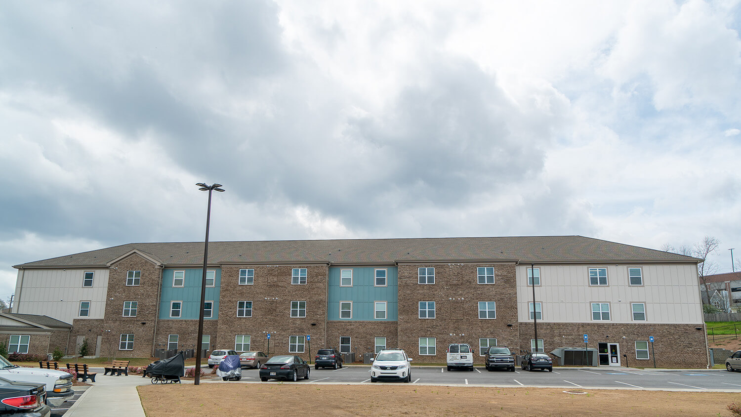 Three-story apartment building with brick and light siding, multiple windows, and front parking lot with cars under a cloudy sky.