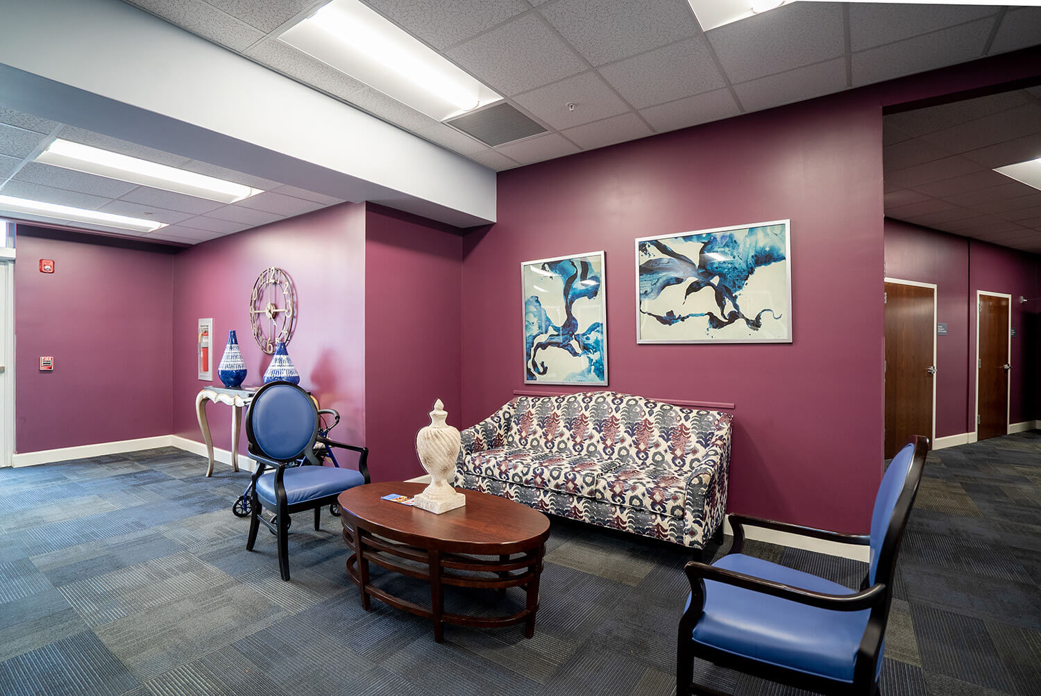 Lobby with purple walls, patterned sofa, two blue chairs, small wooden table, vases, abstract art above sofa, carpeted floor.