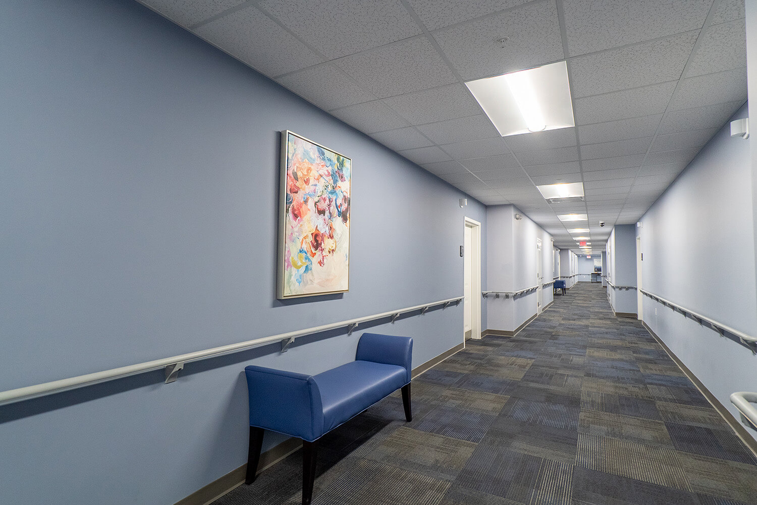 A long, well-lit hallway with light blue walls, blue carpet, handrails, a blue bench, and a colorful abstract painting on the wall. Ceiling lights and doors line the corridor.