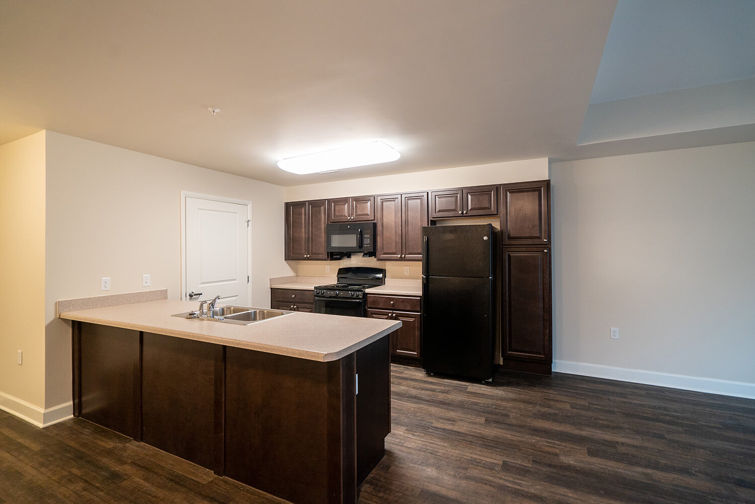 Modern kitchen featuring dark wood cabinets, black appliances, an island with sink, beige counters, and wood flooring under bright lights.