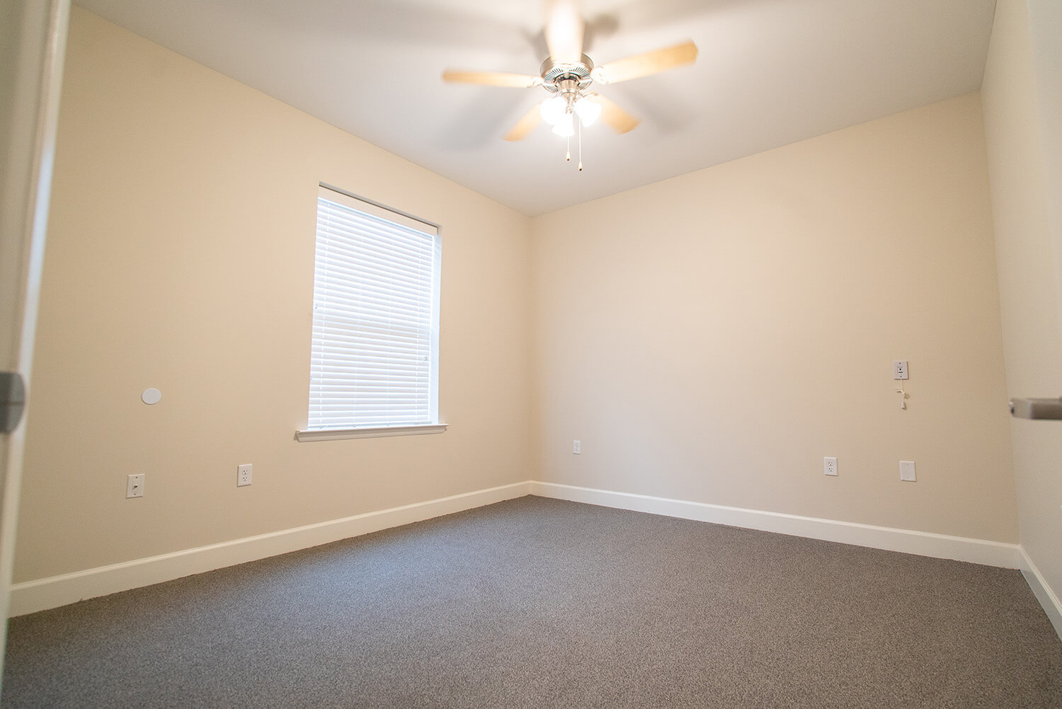 Empty room with beige walls, gray carpet, a window with closed blinds, white trim, ceiling fan with lights, and wall outlets.
