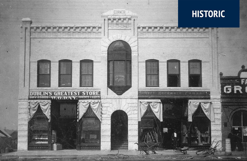Black and white photo of a historic two-story brick building with arched entrance, shops, HISTORIC label, and signs for Dublin's Greatest Store and Gilbert Hardware Company.