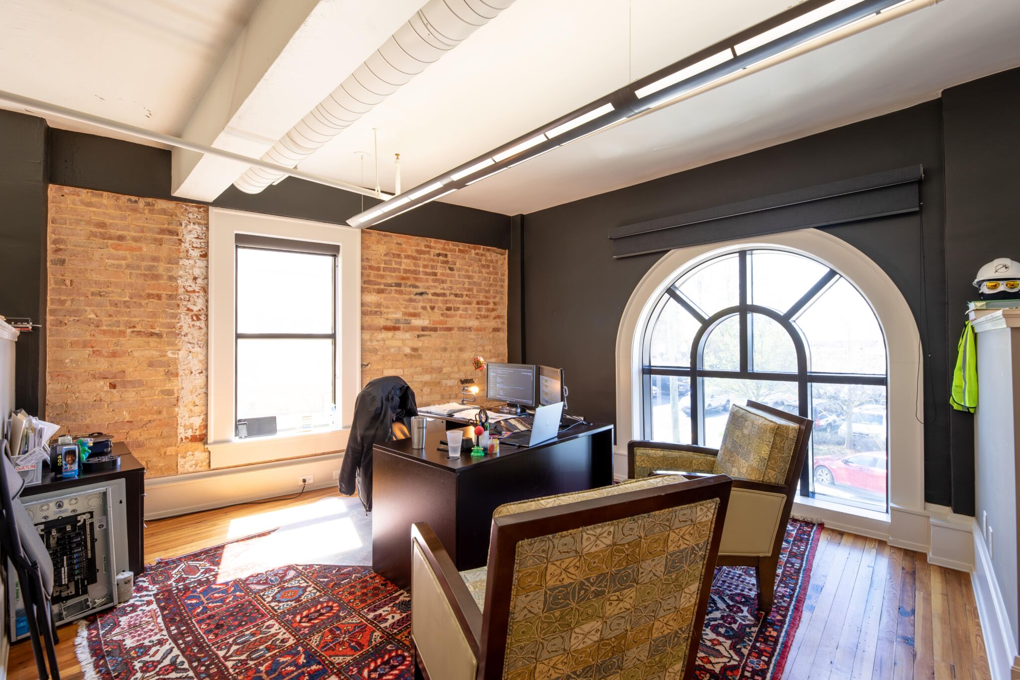 Modern office with exposed brick walls, arched window, wooden desk with monitors, two patterned chairs, colorful rug, natural light.