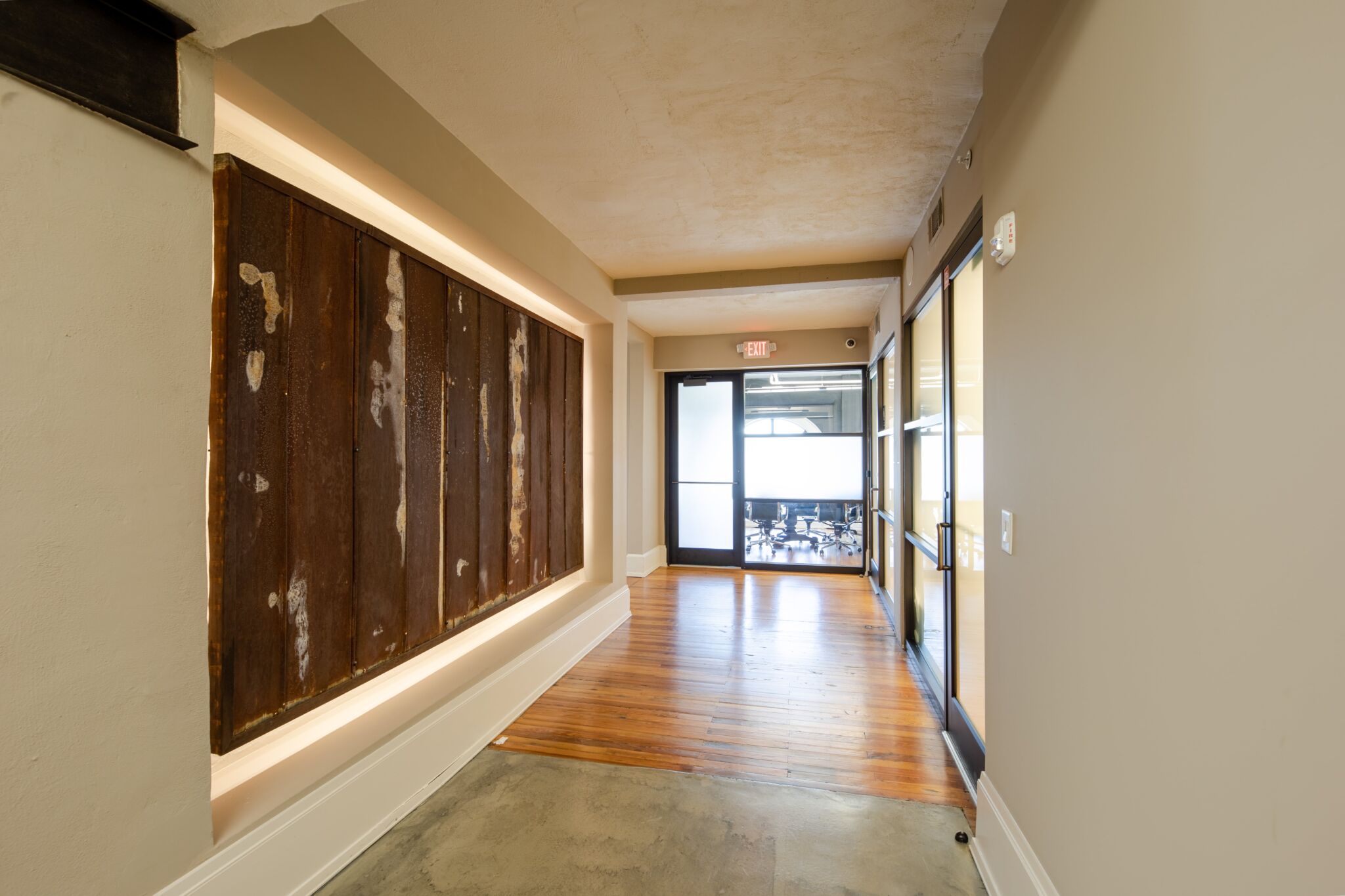 Hallway with beige walls, concrete and wooden floors, large wooden panels left, glass doors at the end to a bright meeting room.