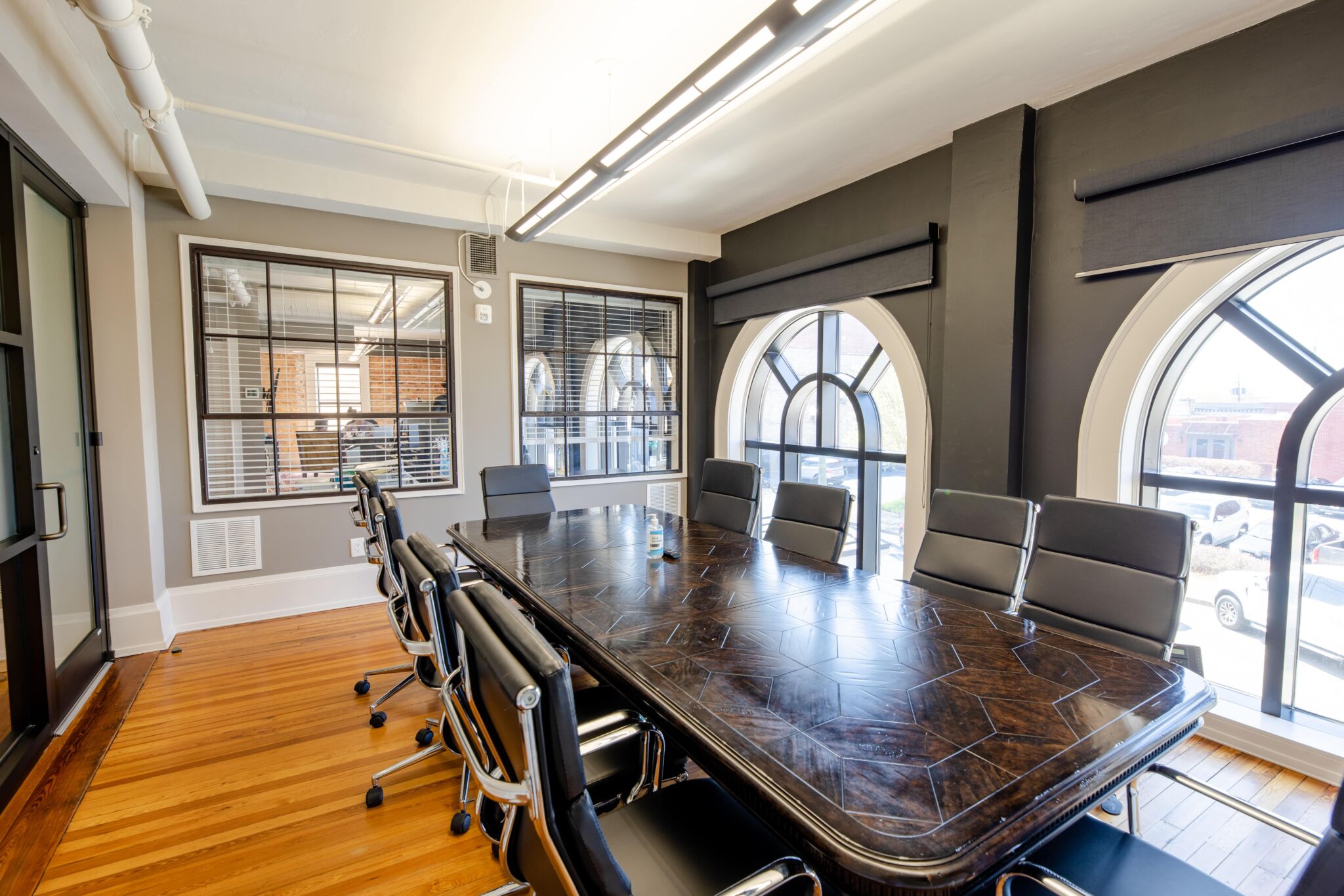 Modern conference room featuring a long dark wood table, black chairs, arched windows, hardwood floors, and glass interior walls.