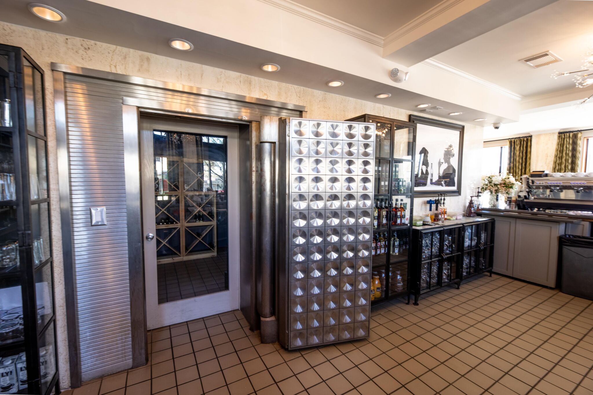 A restaurant interior with a large metallic vault door, shelves of bottles and glassware, tile floors, and art on pale walls.