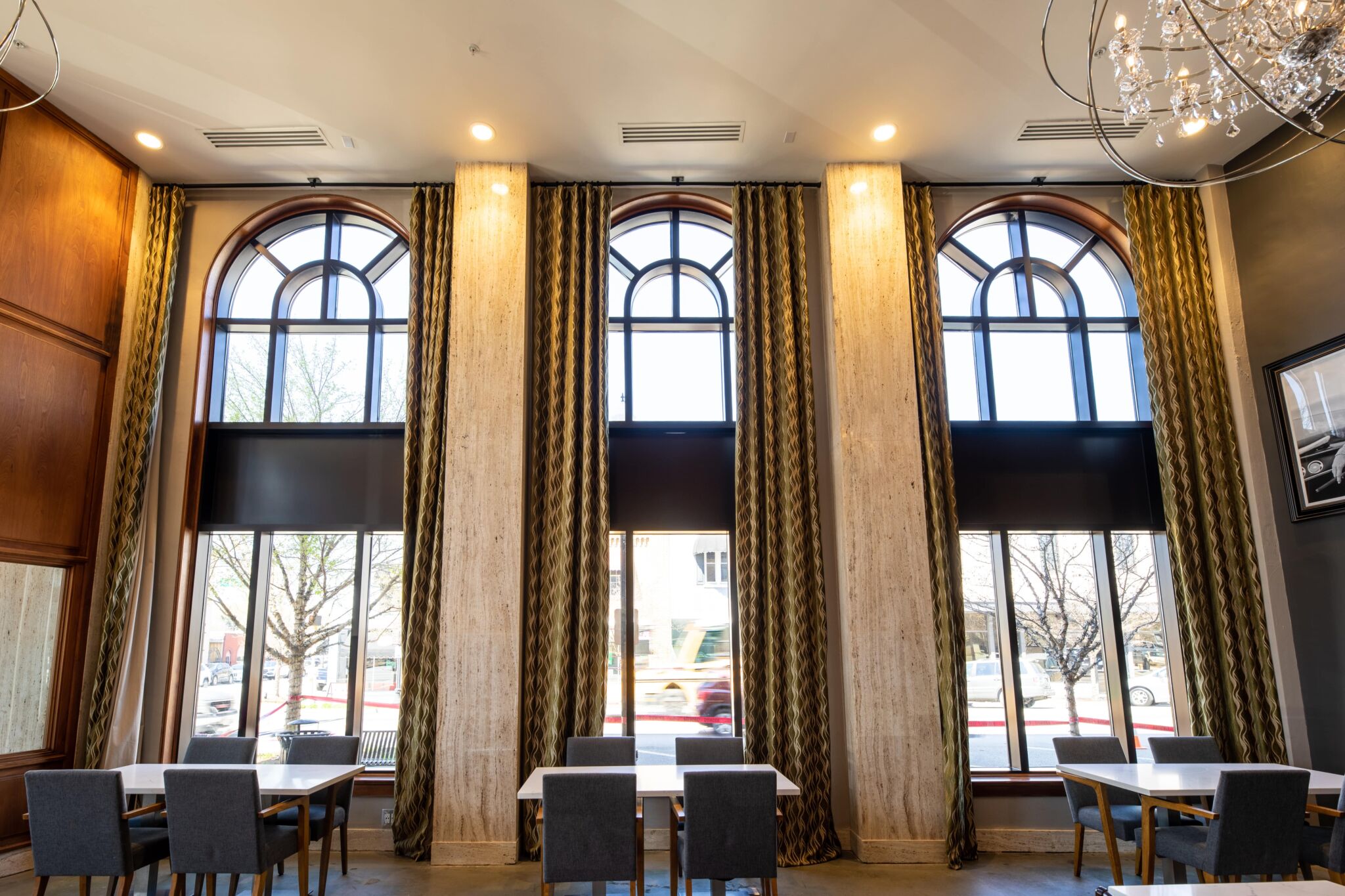 Bright, modern dining area with tall arched windows, patterned curtains, four tables with gray chairs, and trees and cars outside.