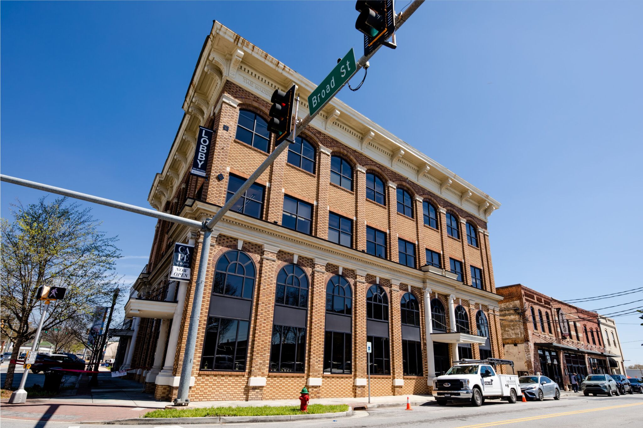 A three-story brick building with large arched windows and a Lobby sign stands at a street corner under a blue sky, with a white pickup truck and Broad St. street sign in the foreground.