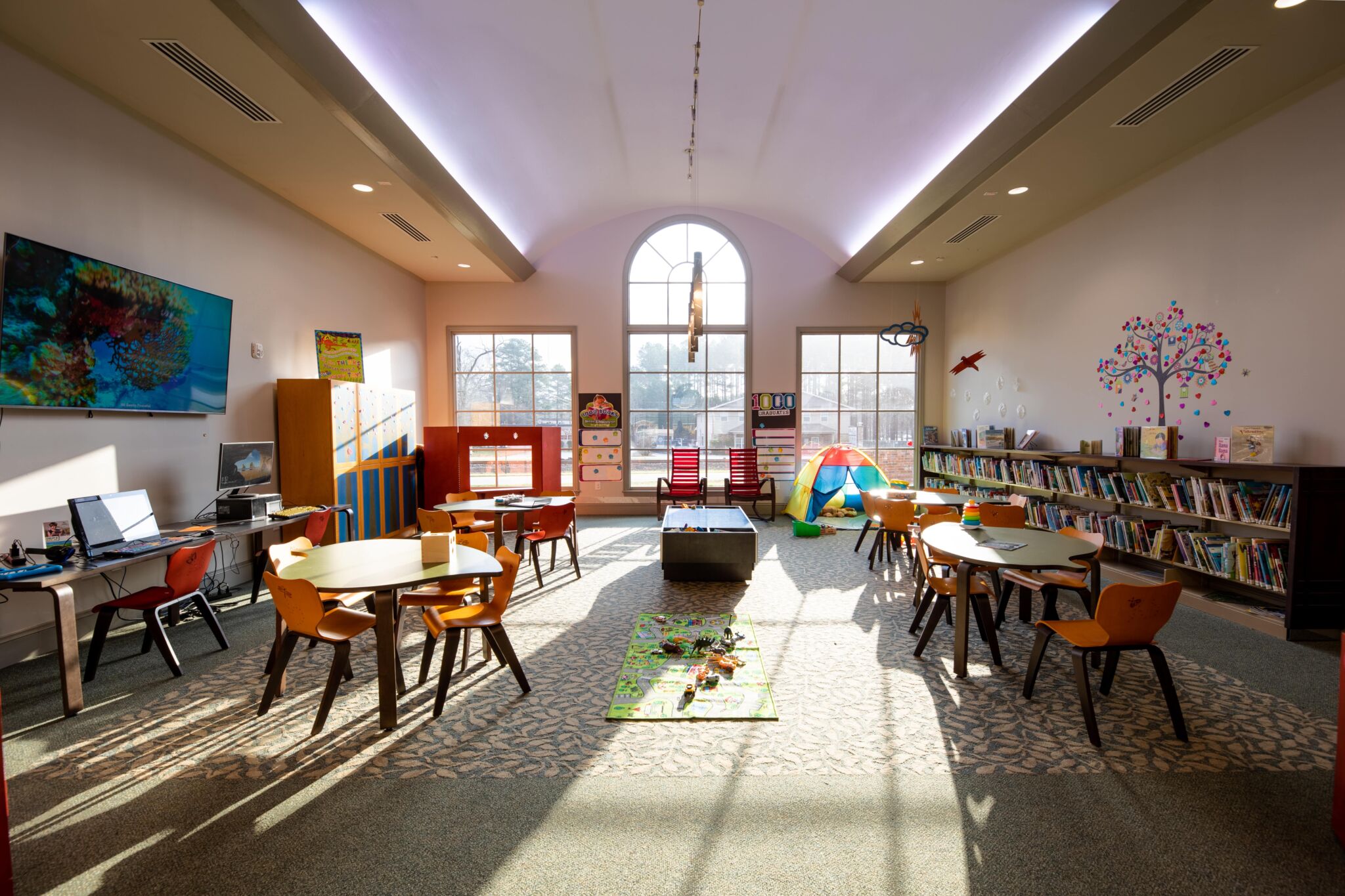 Bright, colorful children's playroom with round tables, chairs, bookshelves, toys, play mat, tent, and sunlight streaming through windows.
