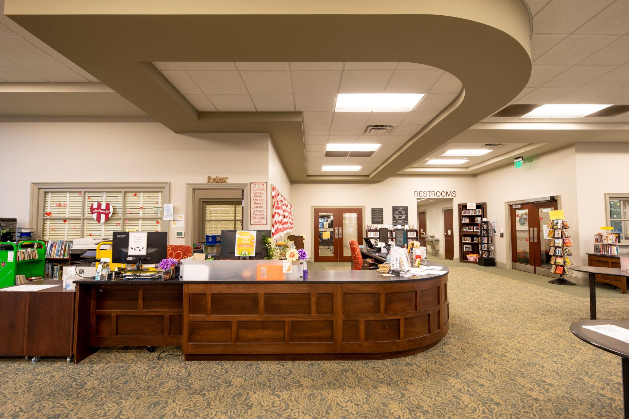 A spacious library interior features a large wooden front desk, bookshelves, and a restroom sign. Ceiling lights brightly illuminate beige carpeted floors.