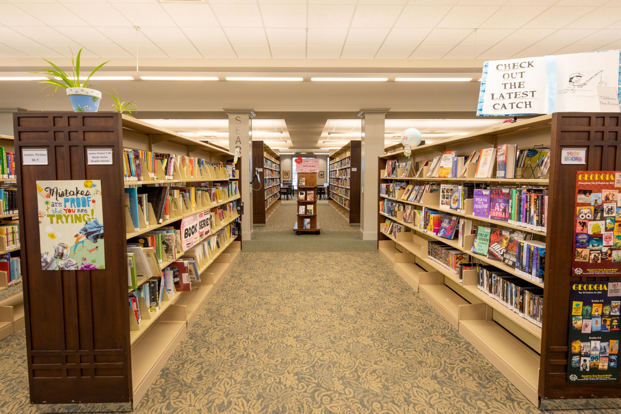 A brightly lit library with tall bookshelves of colorful books on both sides of a carpeted aisle, and a sign reading Check Out the Latest Catch.