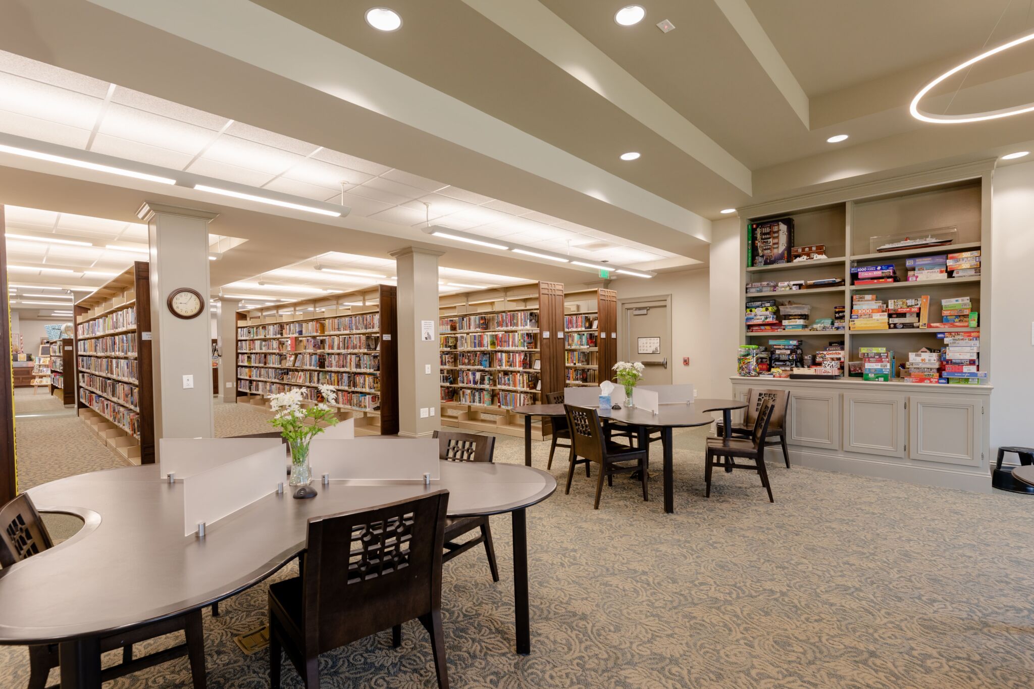 A spacious library with bookshelves, tables and chairs, flowers in vases on tables, a board game shelf, and ceiling lights.