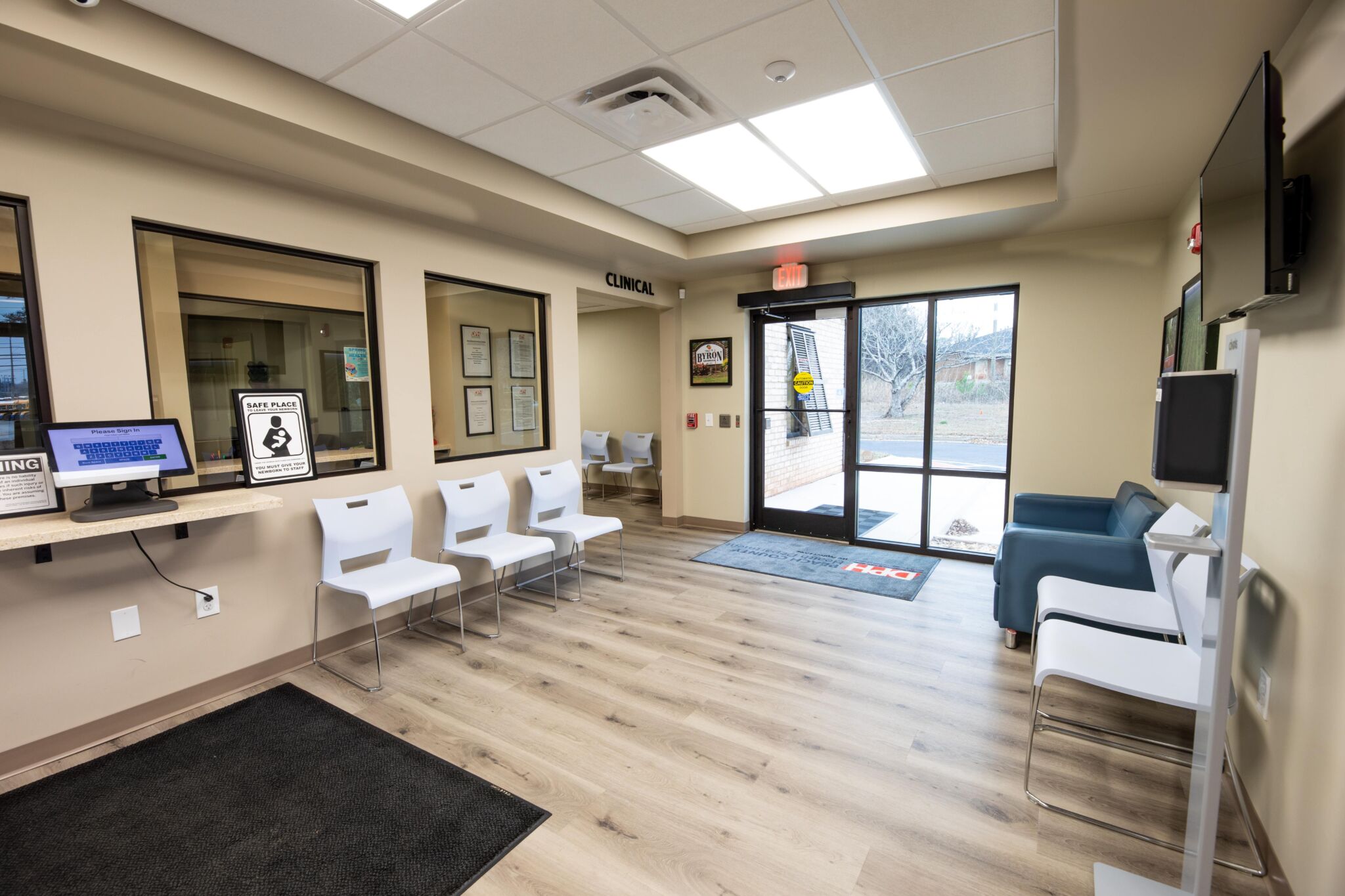 A clean, modern clinic waiting area with white chairs, blue couch, wood floors, big windows, left-side desk, and beige walls.