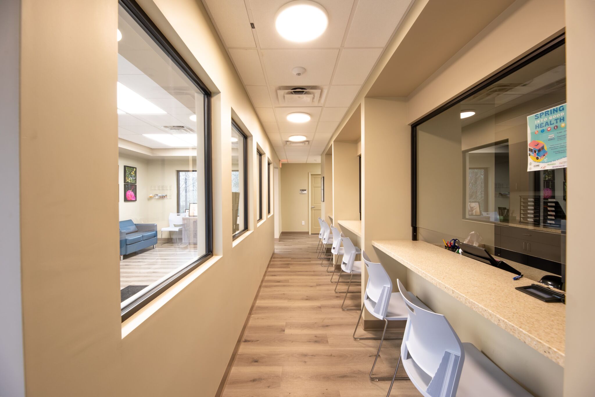 Bright, modern medical office hallway with beige walls, wood flooring, white chairs, large windows to office and waiting area.