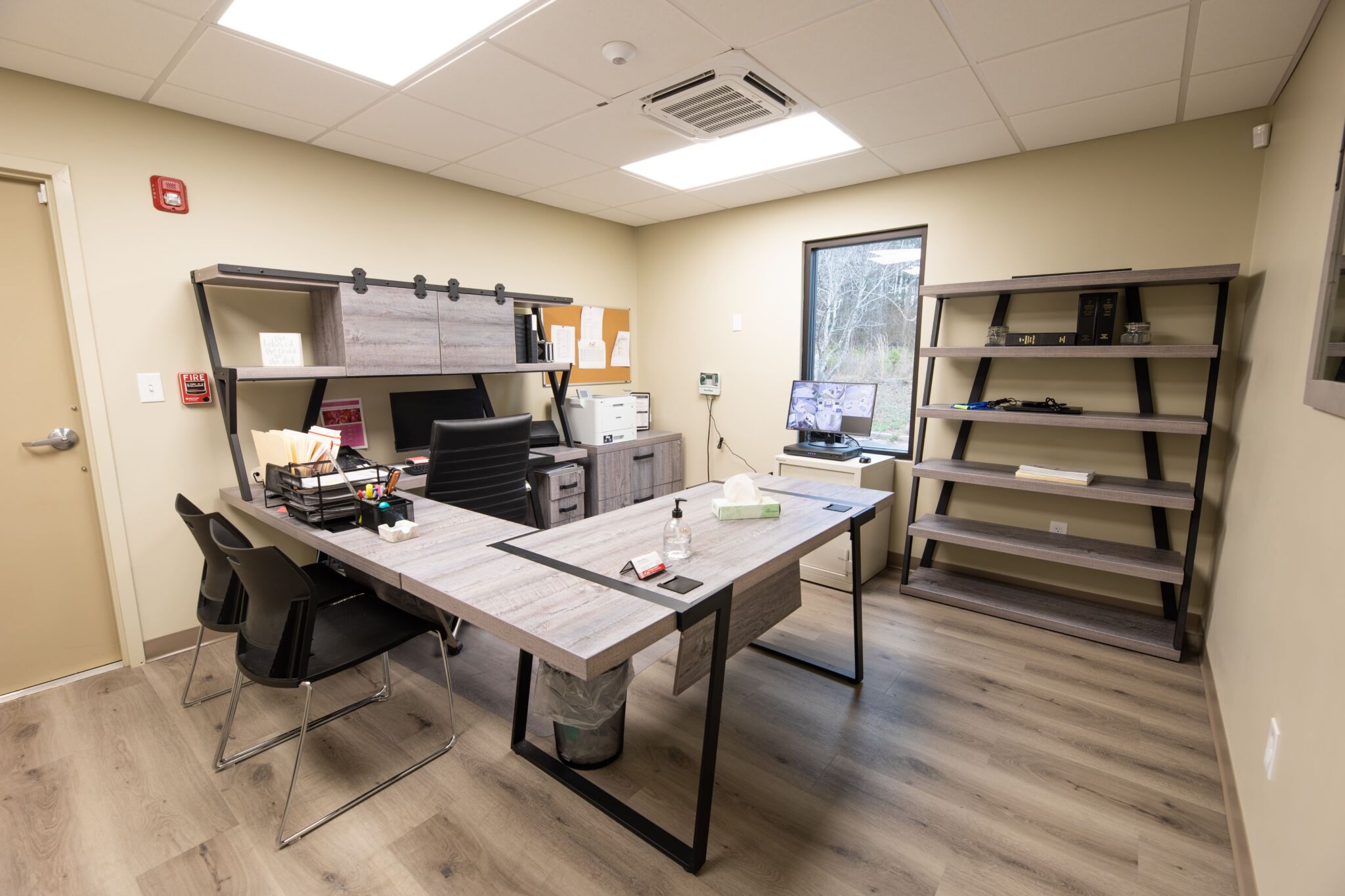 Modern office featuring an L-shaped desk, black chair, guest chairs, monitors, printer, shelves; light wood floor and beige walls.