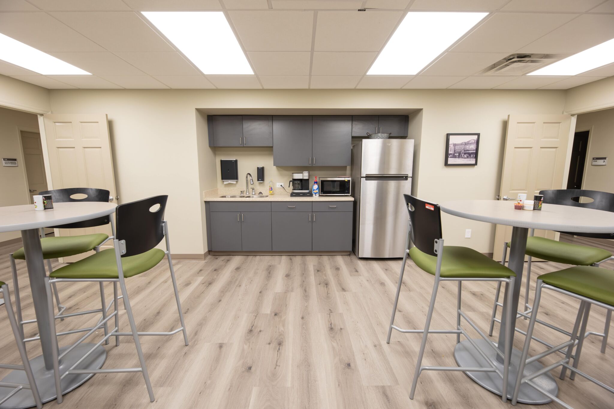 Modern office break room with gray cabinets, stainless steel appliances, and green-cushioned chairs at high tables on wood flooring.
