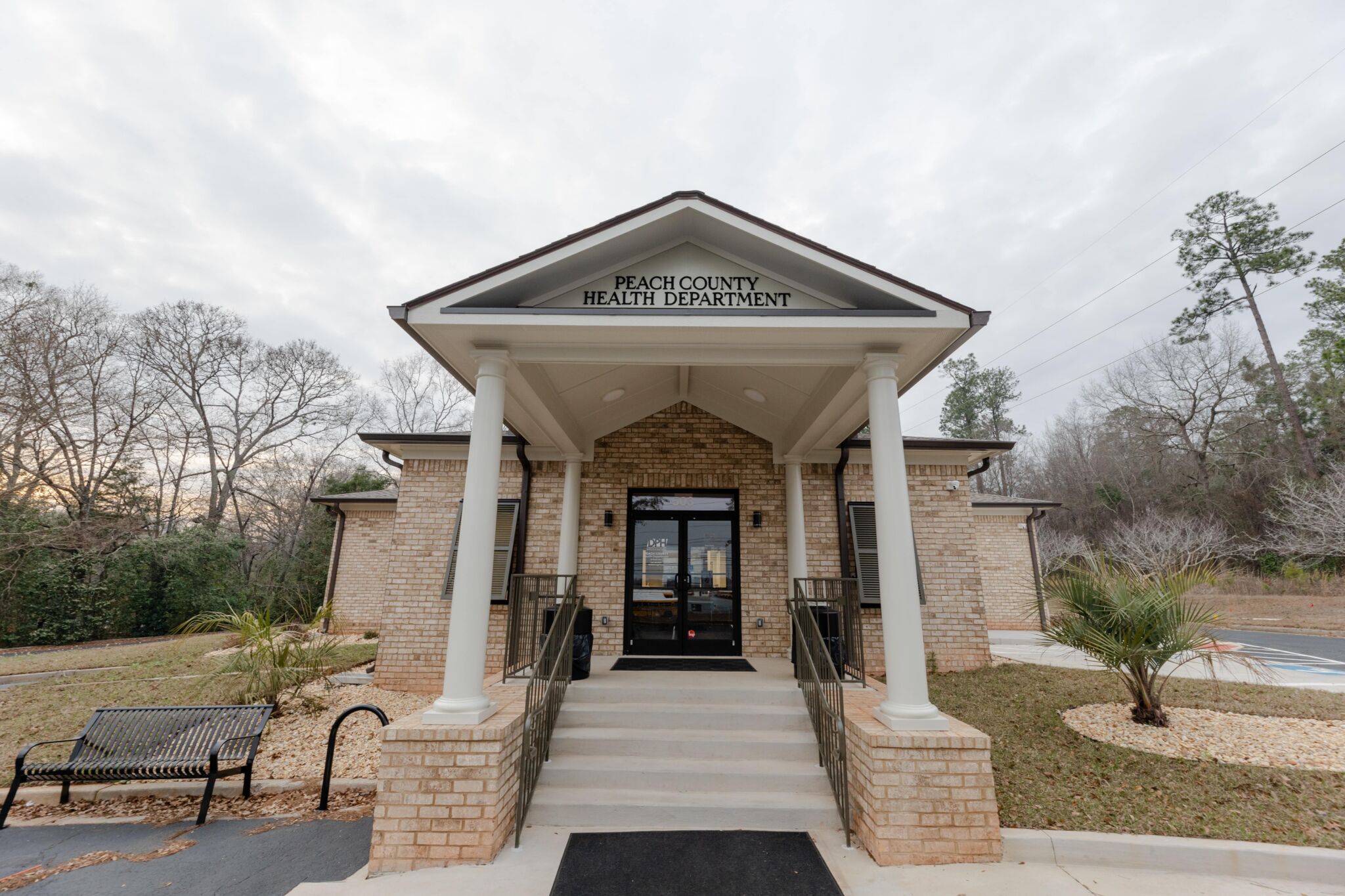 A brick building with a covered entrance labeled Peach County Health Department, pillars, bench, palm trees, and nearby parking lot.