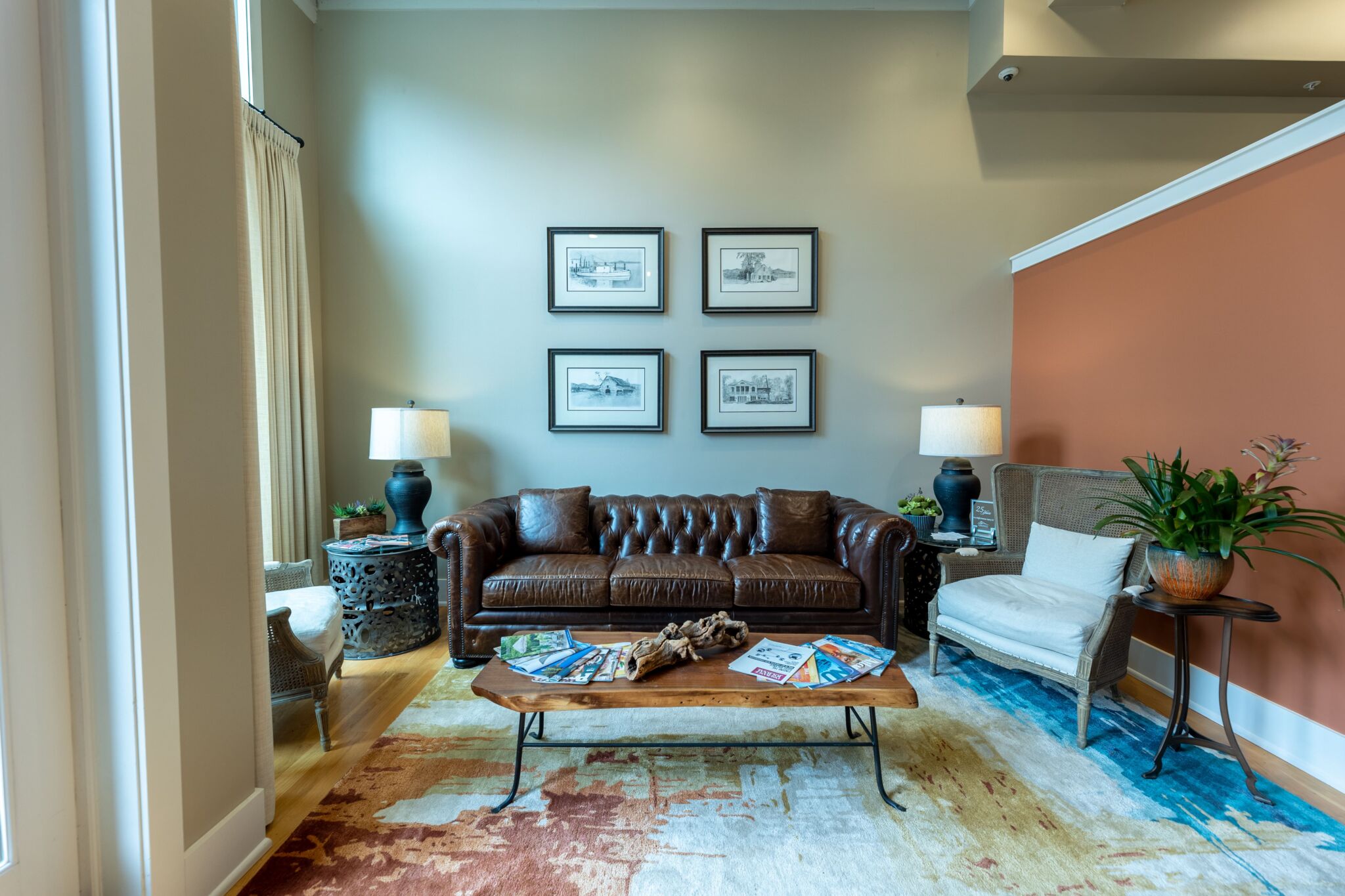 Cozy living room with brown leather sofa, two chairs, wooden coffee table with magazines, table lamps, art, plants, patterned rug.