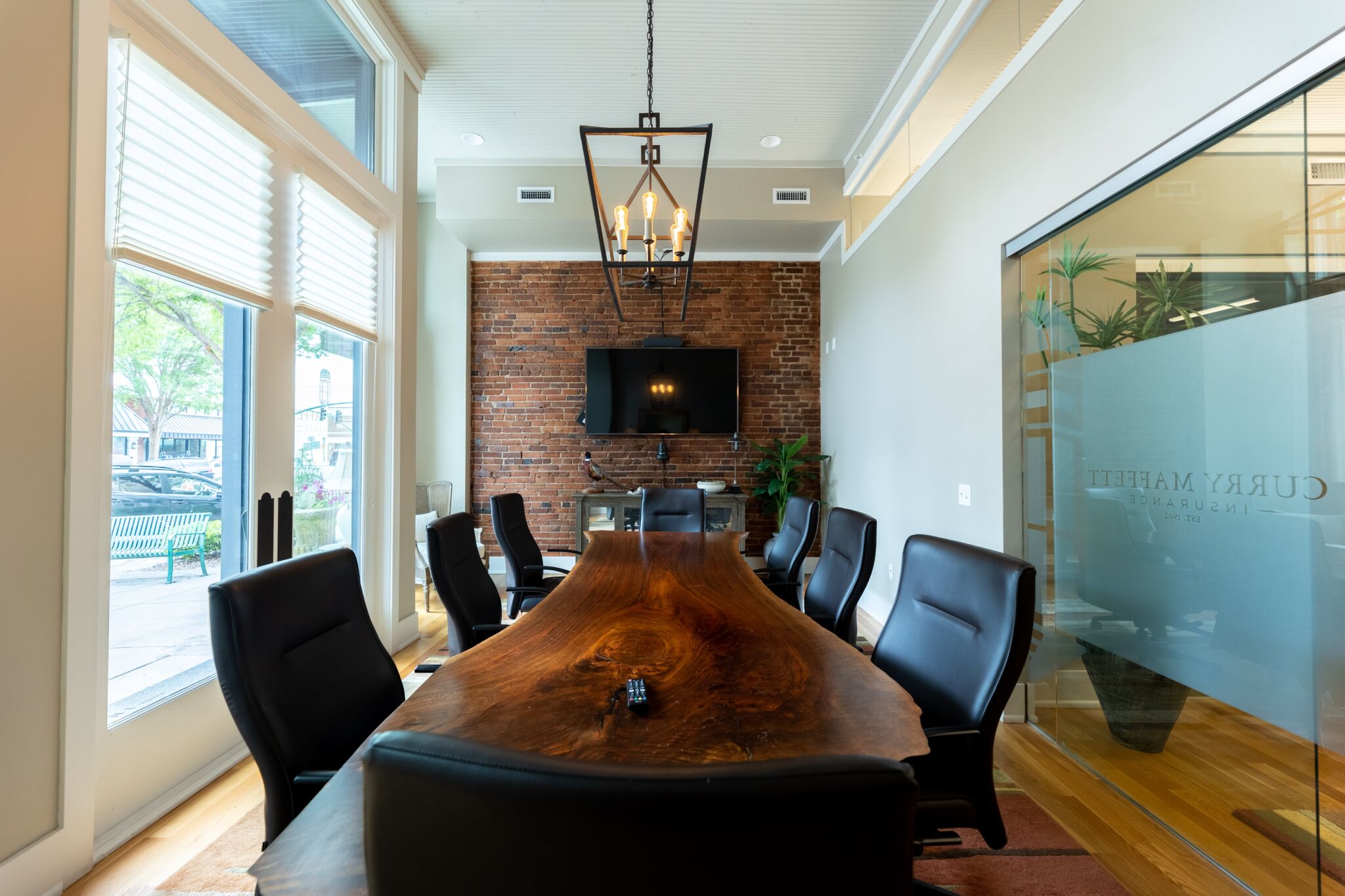 Modern conference room featuring a live-edge wood table, black chairs, TV on exposed brick wall, large windows, and frosted glass walls.