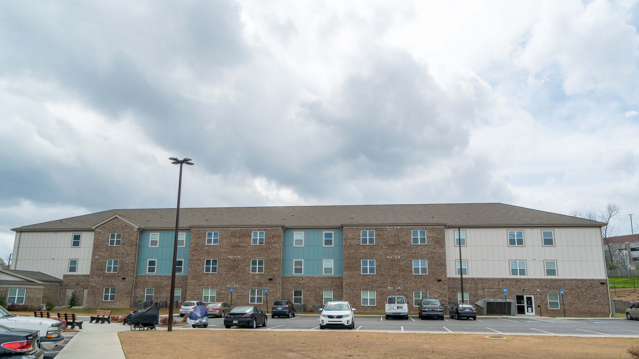 Three-story apartment building featuring brick and light siding, many windows, and a front parking lot with cars under cloudy skies.