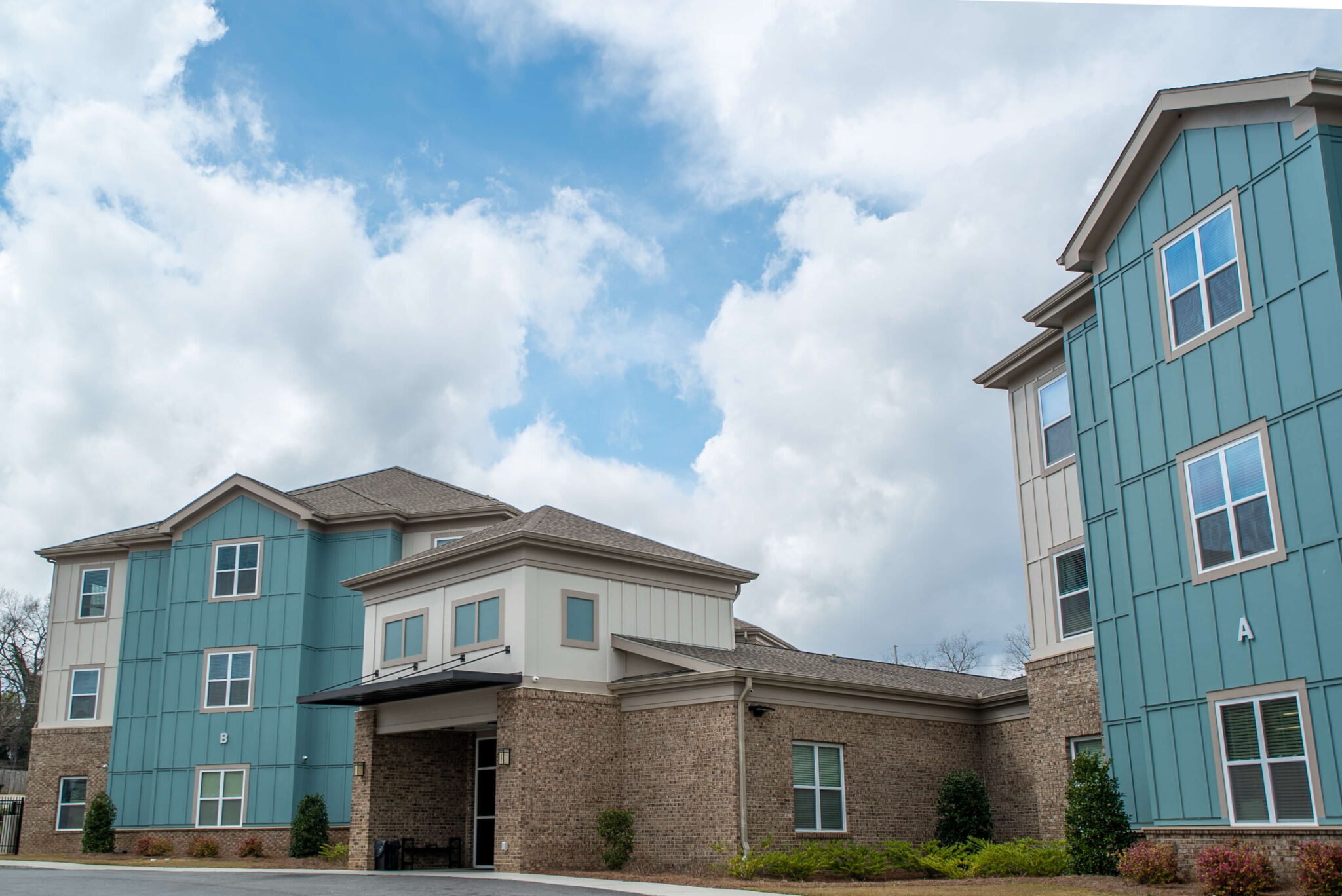 Modern multi-story building with blue and beige siding, brick accents, small bushes around it, beneath a partly cloudy sky.