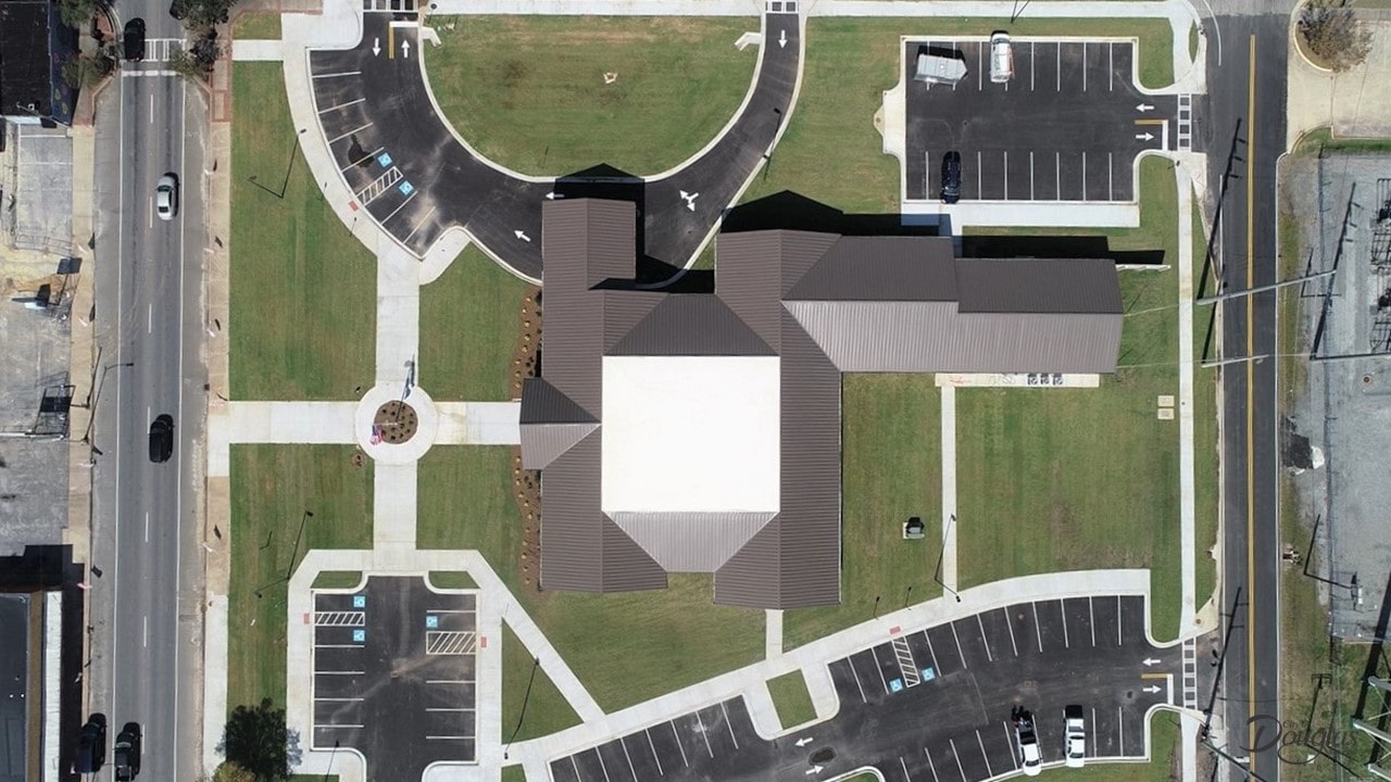 Aerial view of a large building with a white roof, green lawns, sidewalks, and several parking lots at a road intersection.