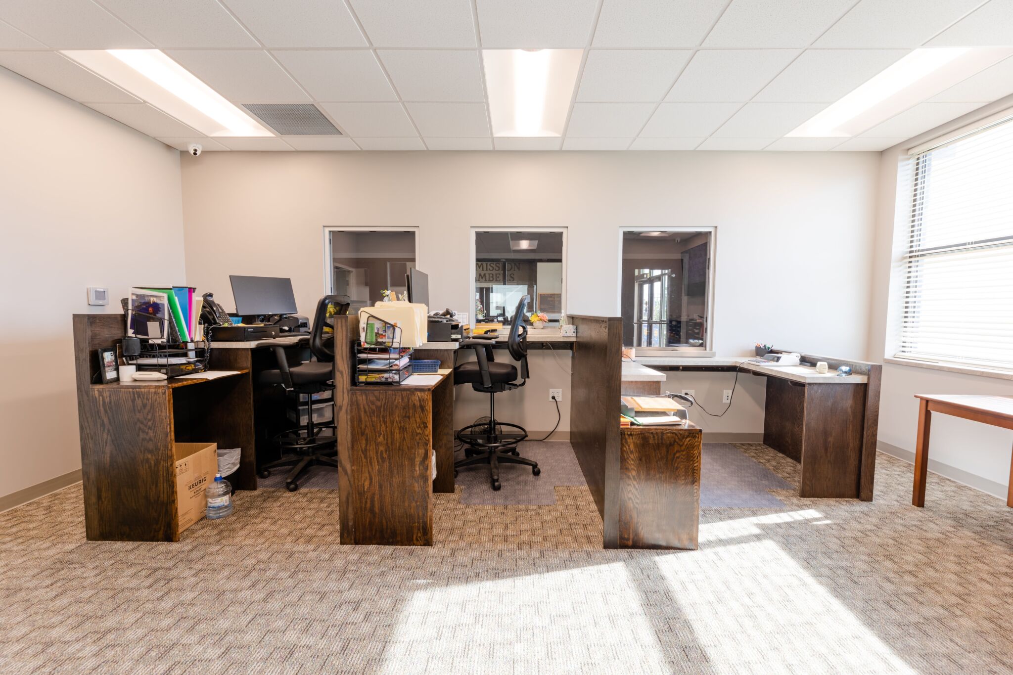A brightly lit office with three wooden desks, glass partitions, computers, supplies, and sunlight streaming in through large windows.